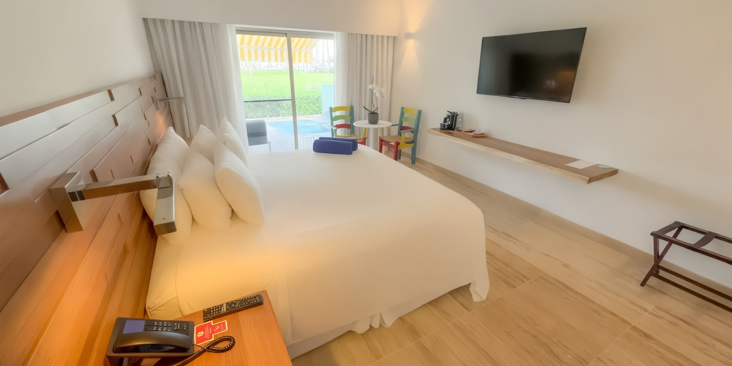 Enjoy a stay at Viva V Samana by Wyndham in a hotel room featuring a spacious bed, a wall-mounted TV, a small table accompanied by chairs, and a sliding door that opens to an outdoor area.