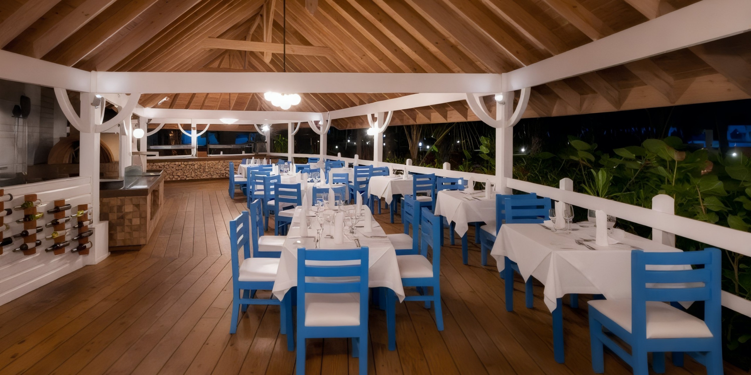 Viva V Samana by Wyndham offers an outdoor dining area featuring wooden floors and blue chairs surrounding tables draped with white cloths. Wine bottles are showcased on display, with a chandelier elegantly hanging overhead, and lush greenery visible beyond the dining space.