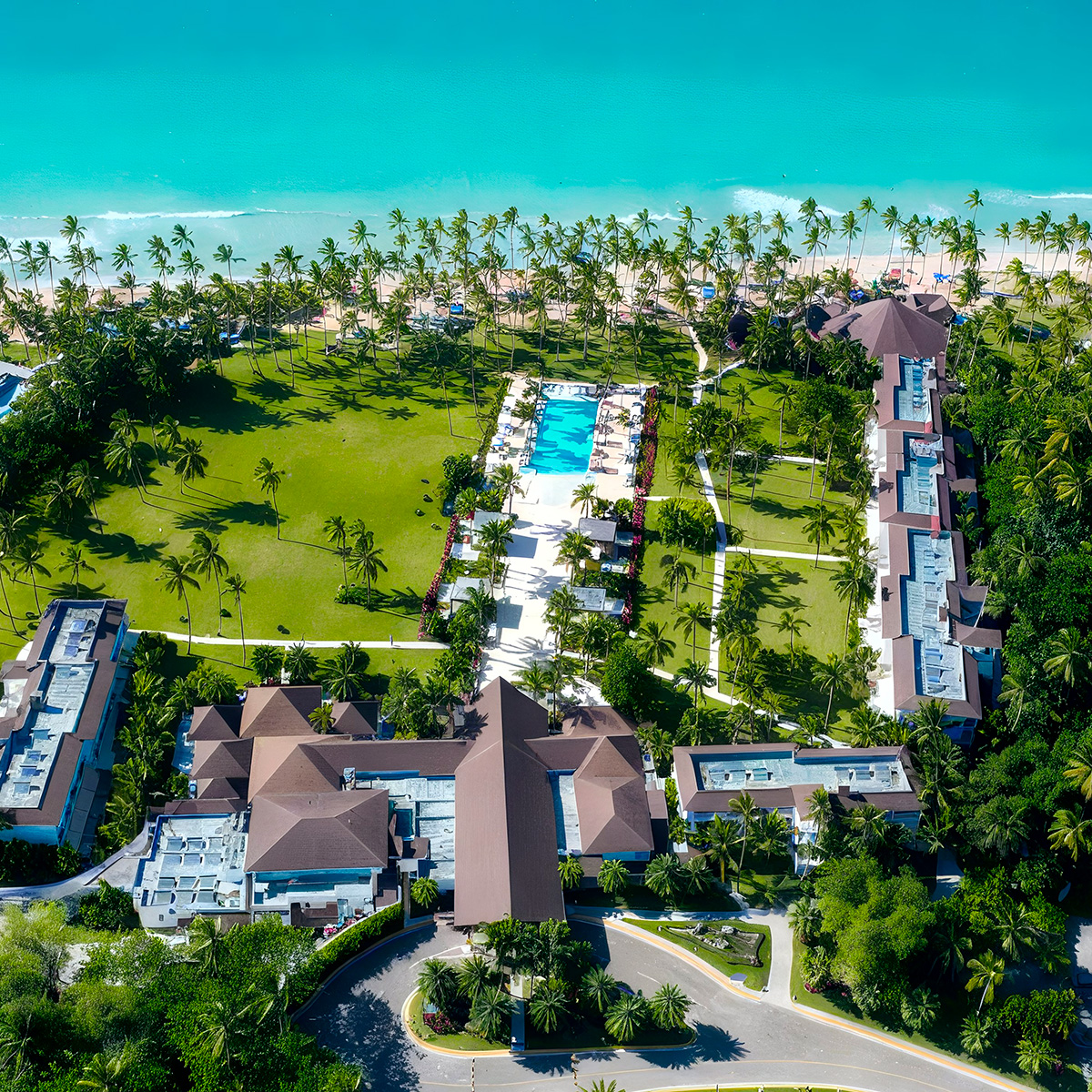 An aerial view of Viva V Samana by Wyndham, a beachfront resort with a central pool, surrounded by green lawns, palm trees, and multiple buildings, set against the backdrop of a turquoise ocean.