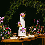 A tiered wedding cake adorned with purple and pink flowers is surrounded by floral arrangements and trays of pastries on a wooden table.