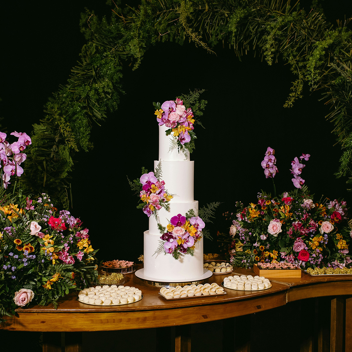 A tiered wedding cake adorned with purple and pink flowers is surrounded by floral arrangements and trays of pastries on a wooden table.