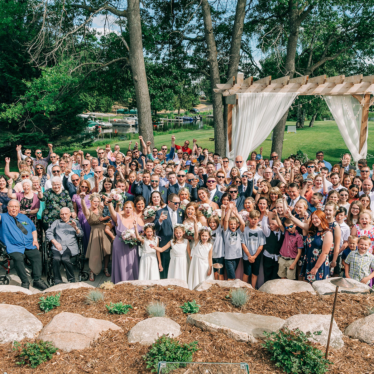 Large outdoor wedding gathering with guests of all ages posing together under trees and a wooden pergola.