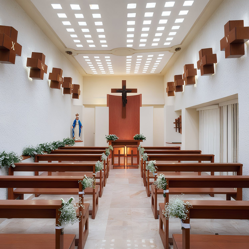 A small, modern chapel with wooden benches, a crucifix on the wall, decorative plants on the benches, and a statue of Mary in the corner.