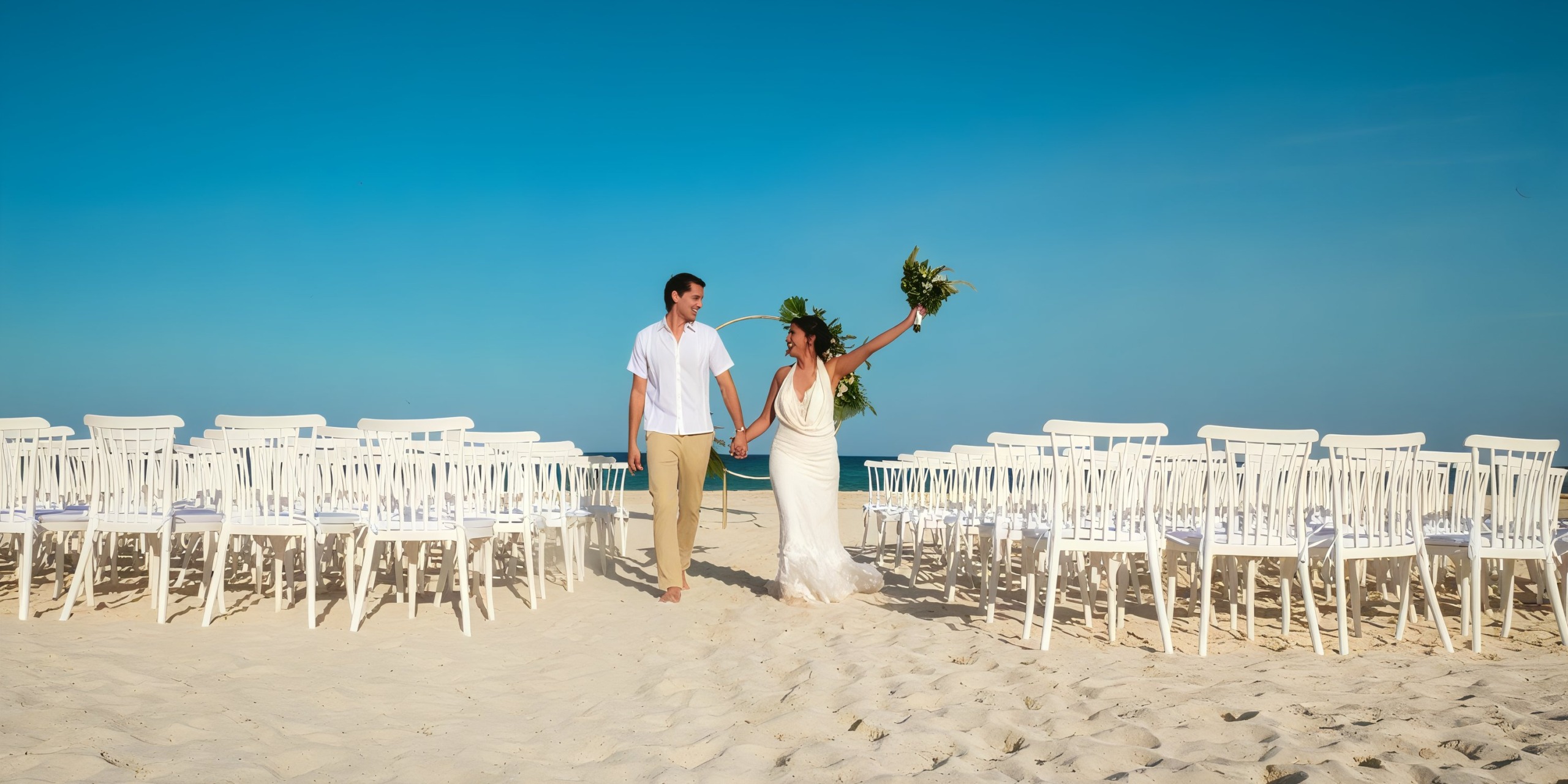 A couple in wedding attire holding hands and walking on a sandy beach aisle between rows of white chairs at Wyndham Grand Cancun, with a clear blue sky in the background.