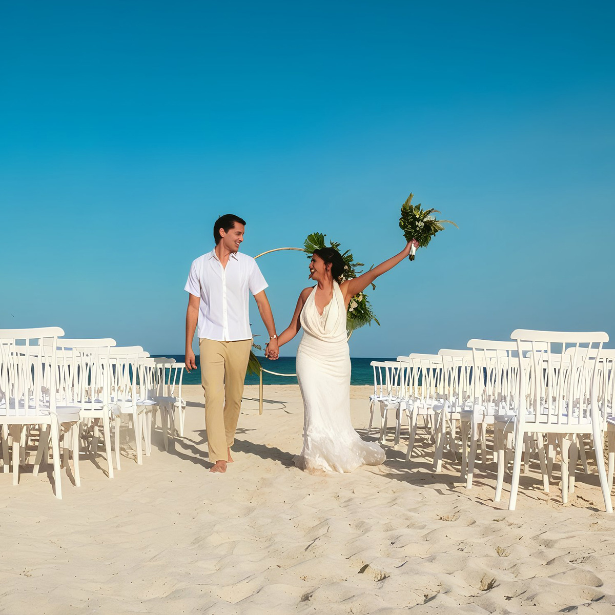 A couple holding hands, walking between white chairs on a sandy beach, with the ocean in the background. The woman holds a bouquet, and both are smiling under a clear blue sky.
