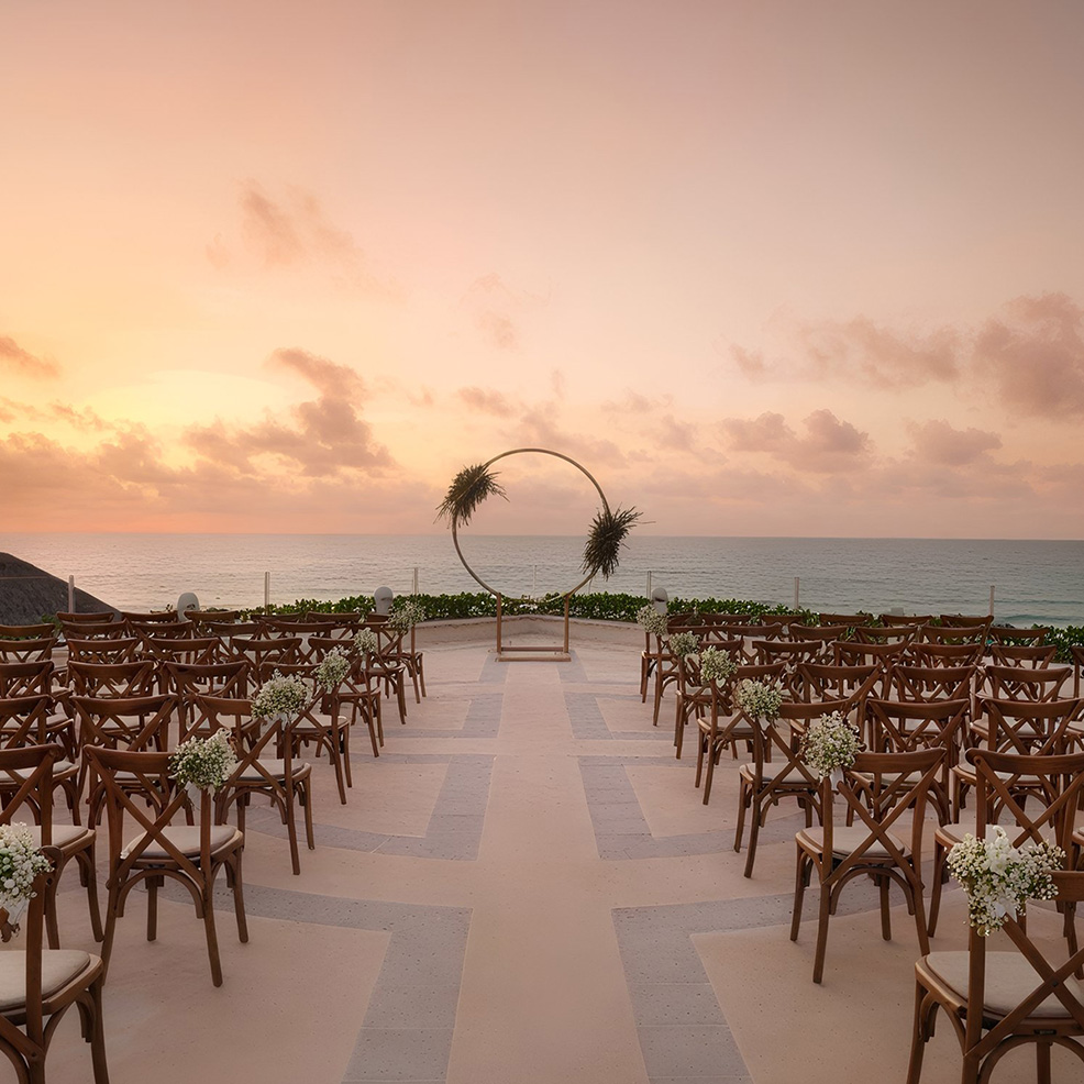 Outdoor wedding setup with wooden chairs arranged in rows facing a circular arch adorned with greenery, overlooking the ocean at sunset.