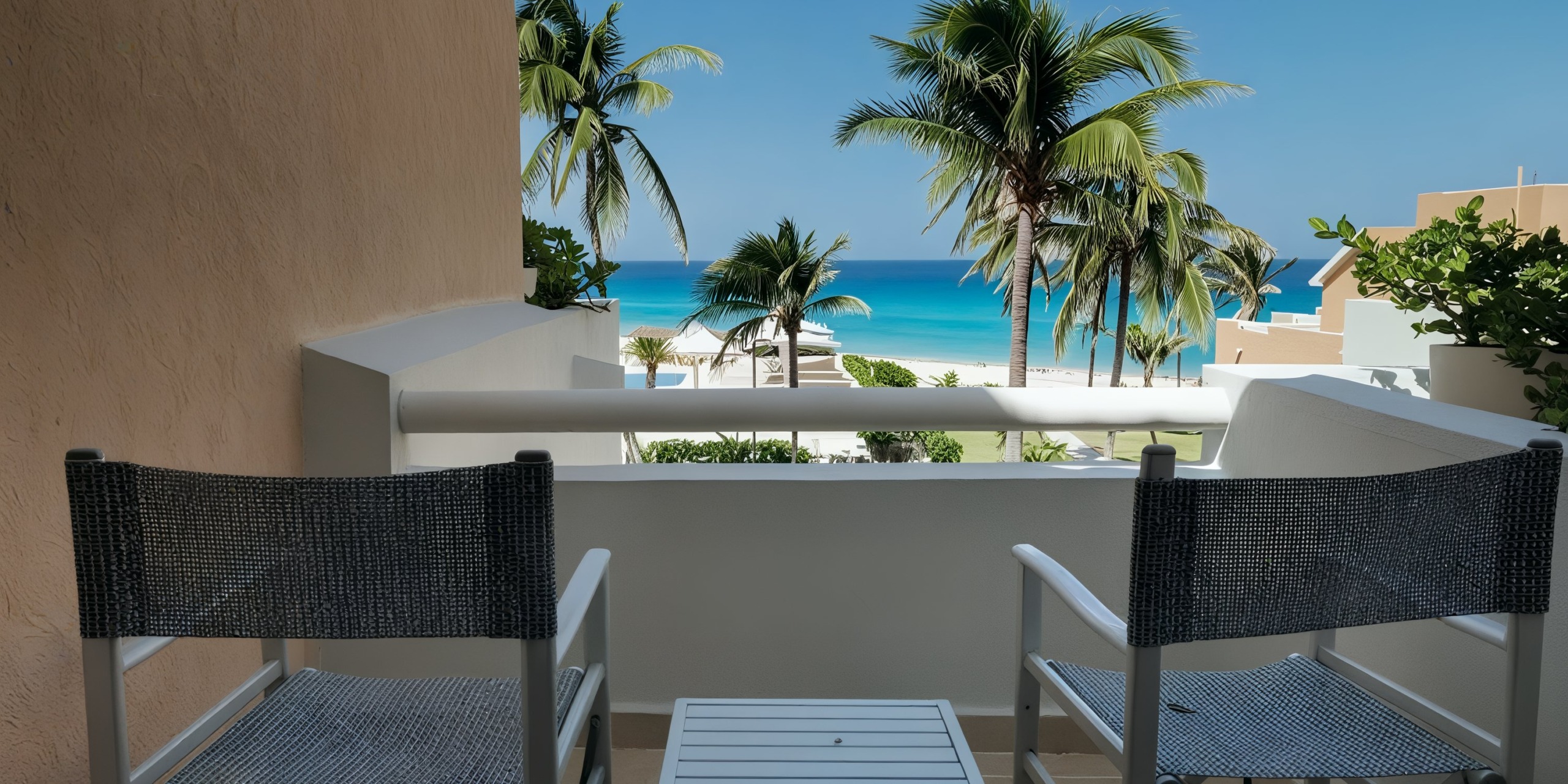 On the balcony of the Wyndham Grand Cancun, two chairs and a table offer views of palm trees, a sandy beach, and the sparkling blue ocean beneath a clear sky.