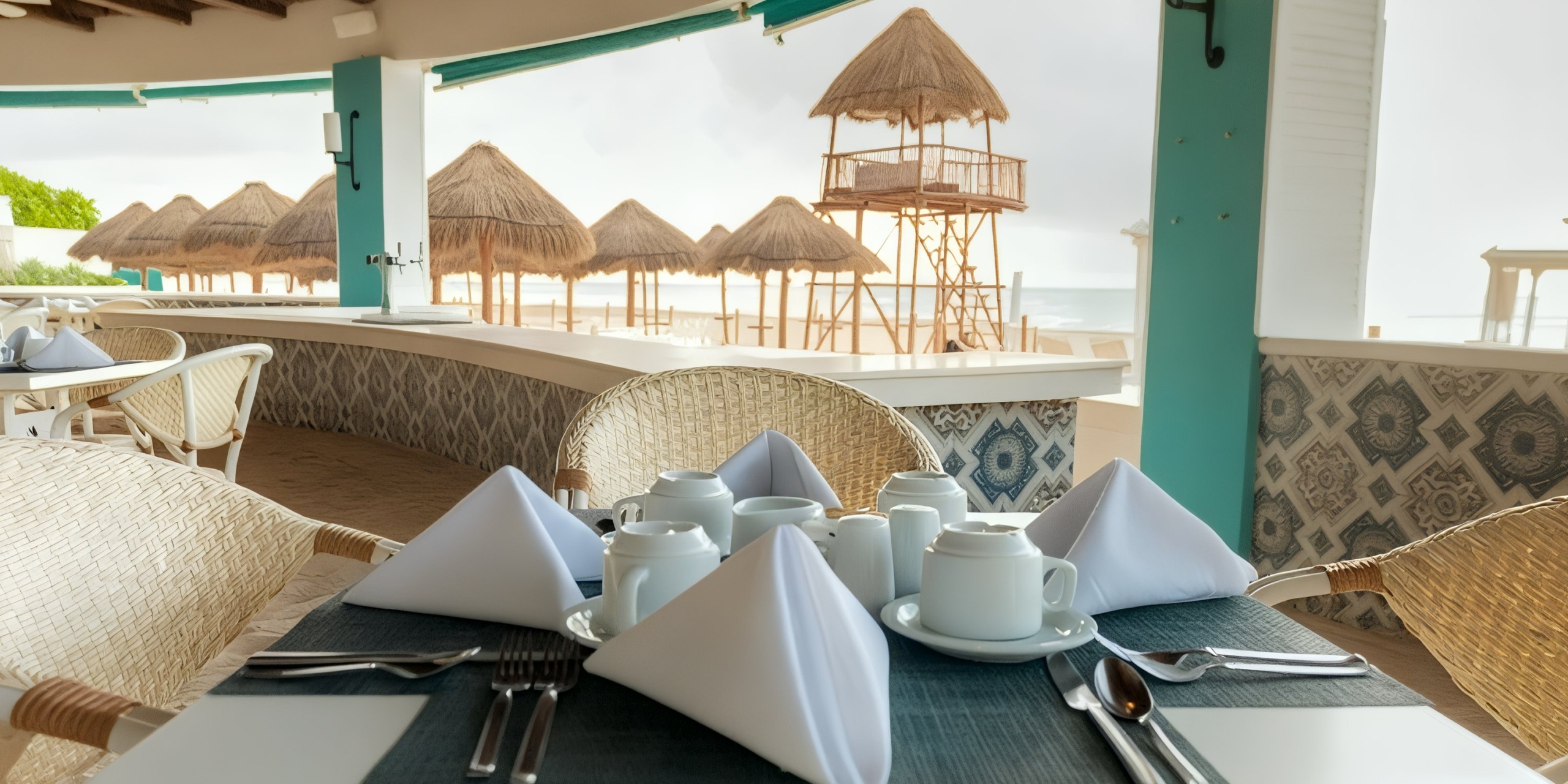 Enjoy breakfast in the outdoor dining area at Wyndham Grand Cancun, with a table set featuring white napkins and cups. The view overlooks a sandy beach adorned with thatched umbrellas and a lifeguard tower in the background.