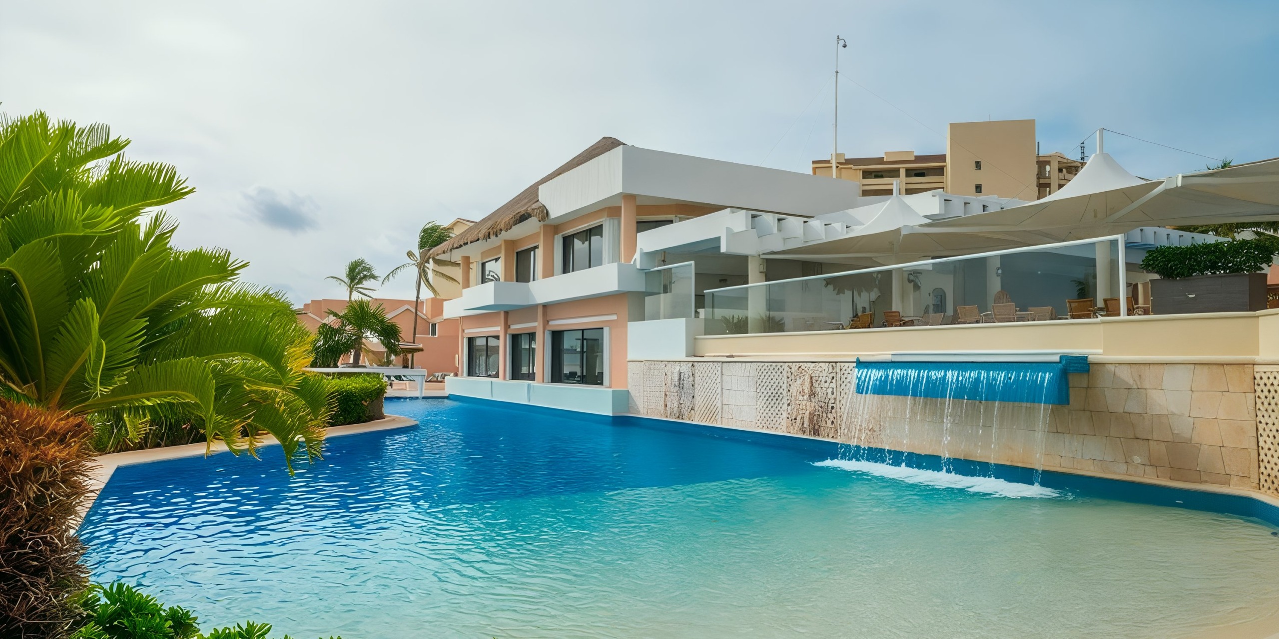 The modern exterior of the Wyndham Grand Cancun features a large pool and a waterfall, all surrounded by palm trees. In the background, white and beige buildings stand under a partly cloudy sky.