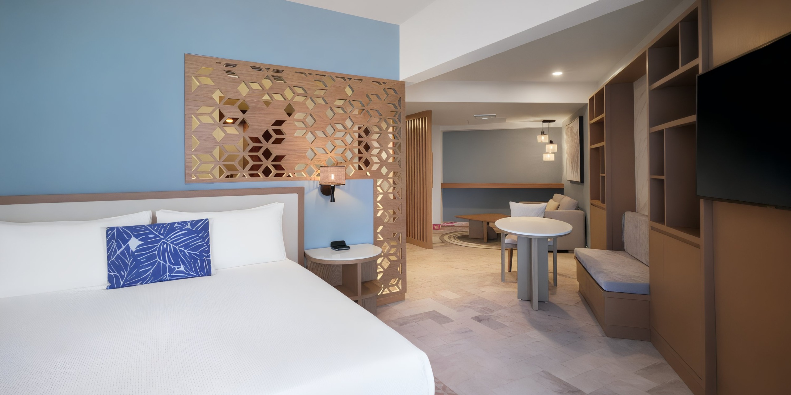 Experience the contemporary elegance of a Wyndham Grand Cancun room, featuring a king-size bed, decorative partition, a cozy table with chairs, light blue walls complemented by wooden accents, abundant natural light, and a sleek design.