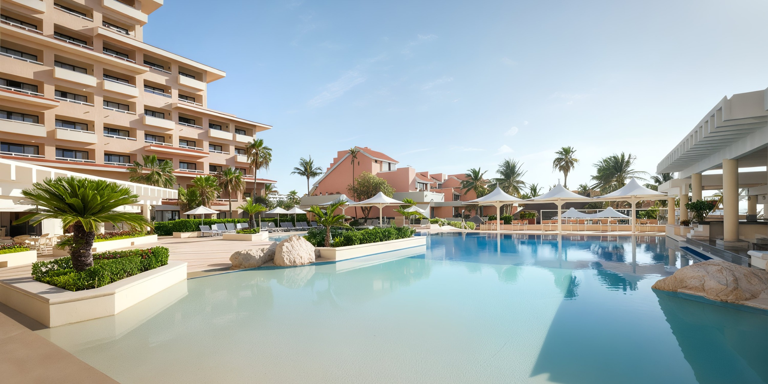 Wyndham Grand Cancun's resort pool is adorned with lounge chairs and umbrellas, encircled by swaying palm trees and nearby buildings, all set beneath a clear sky.