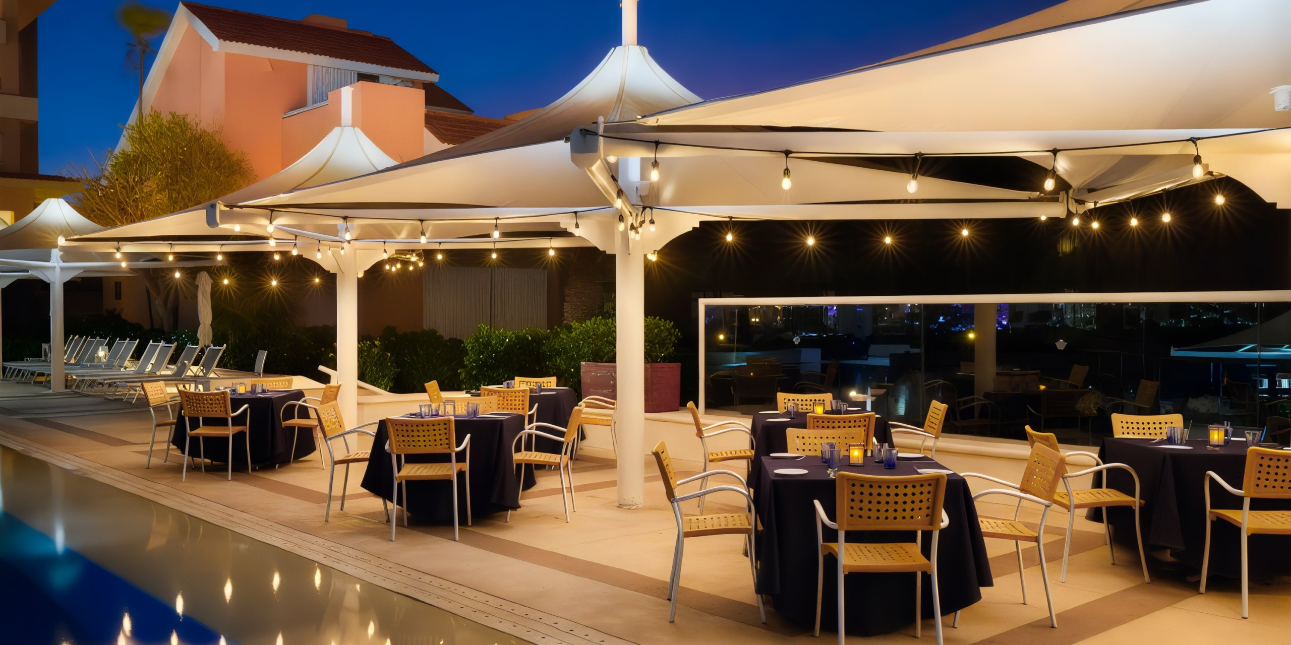 Dine outdoors at the Wyndham Grand Cancun, with tables and chairs set under illuminated canopies beside the pool in the evening.