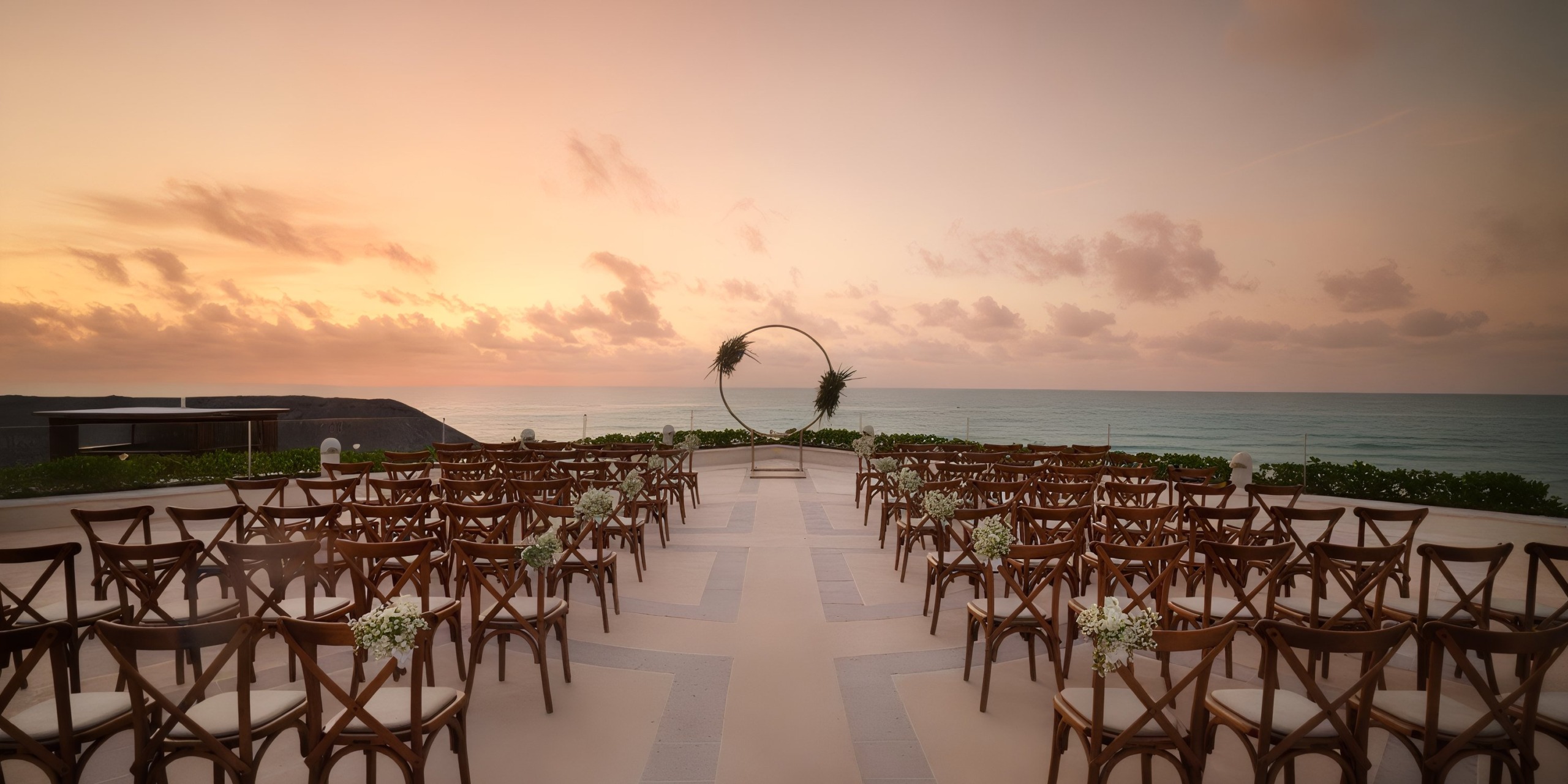 Create an outdoor wedding setup at the Wyndham Grand Cancun with wooden chairs arranged to face a circular arch adorned with greenery, and enjoy the breathtaking ocean view as the sun sets.