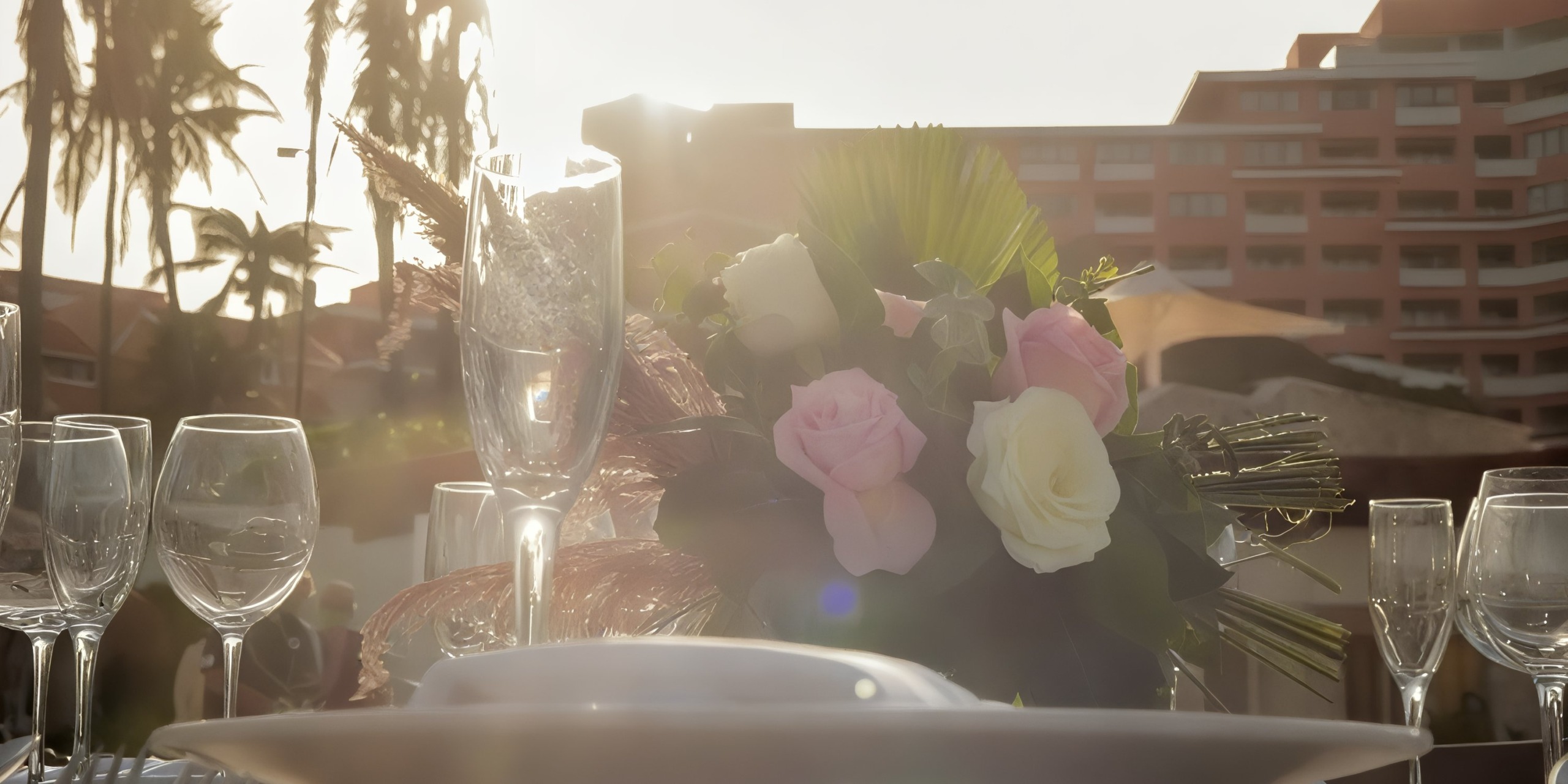 The Wyndham Grand Cancun serves as the backdrop for a beautifully arranged outdoor table set with champagne glasses and a floral centerpiece featuring pink and white roses, all illuminated by the setting sun near palm trees.