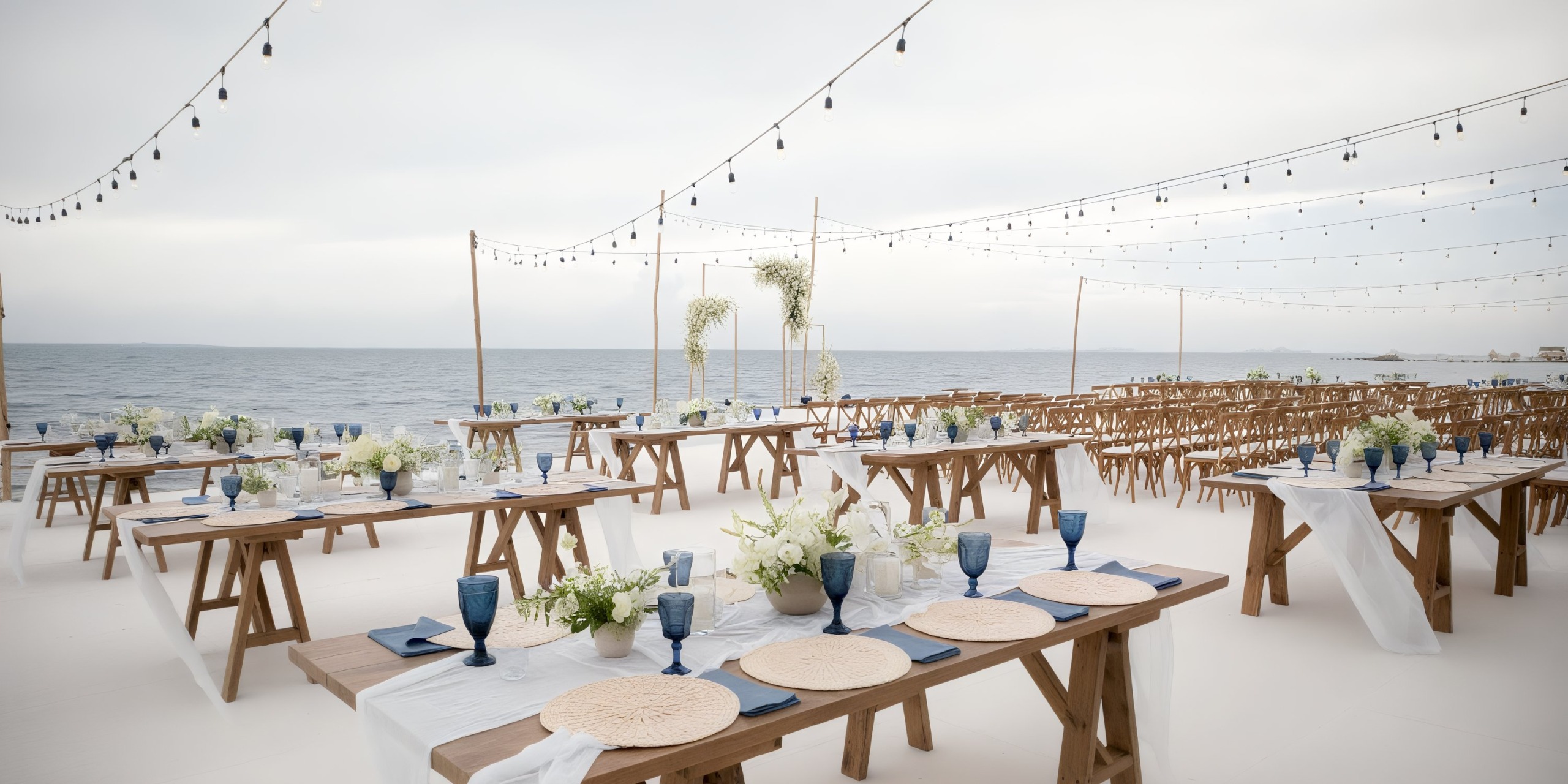 Outdoor wedding reception setup at Garza Blanca Cancun, featuring wooden tables and chairs, blue glasses, and string lights overhead by the sea.