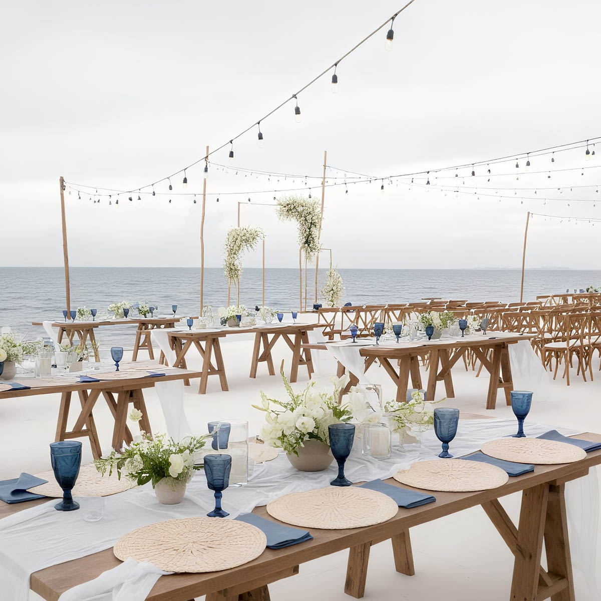Outdoor beach wedding setup with wooden tables and chairs, blue glassware, white flowers, beige placemats, and string lights overhead against an ocean backdrop.