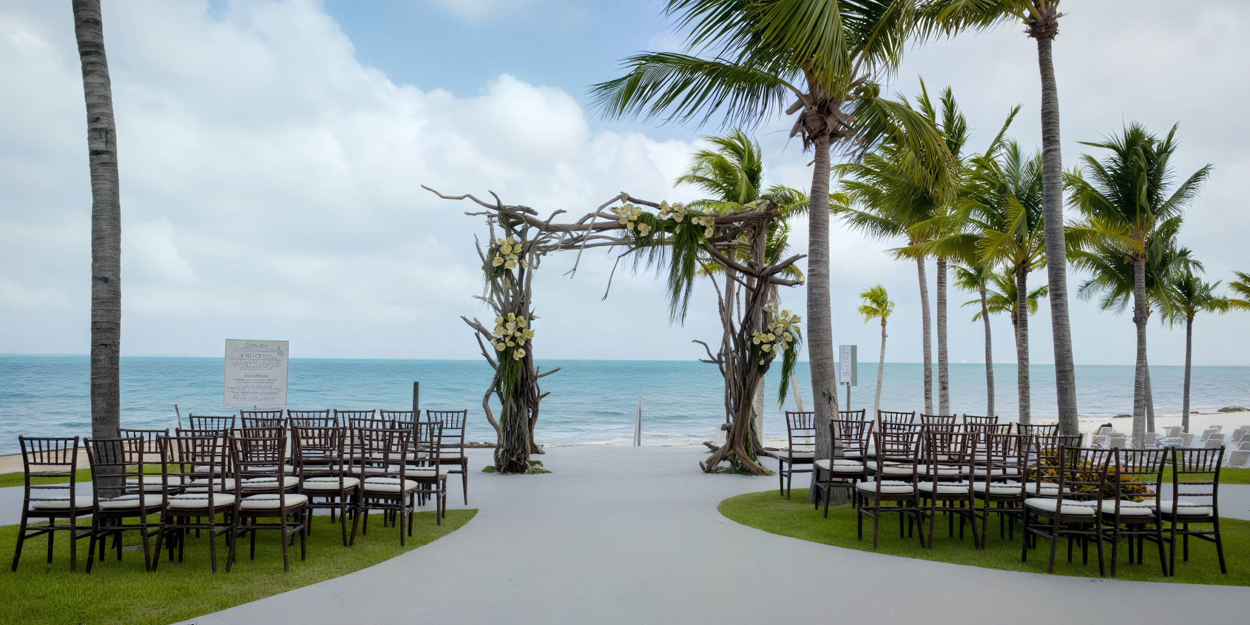 Garza Blanca Cancun offers a beachfront wedding setup featuring a wooden arch adorned with flowers, white guest chairs, palm trees, and an ocean view.