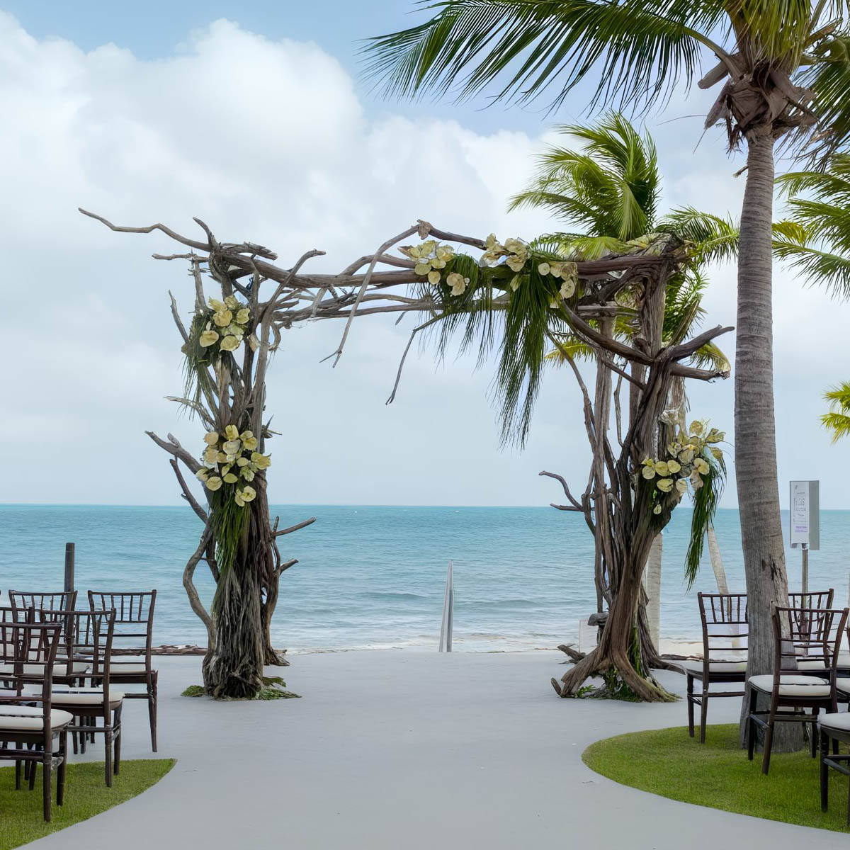 A beachside wedding arch made of driftwood and adorned with white flowers and palm fronds stands near rows of empty chairs. The ocean and palm trees are in the background under a partly cloudy sky.