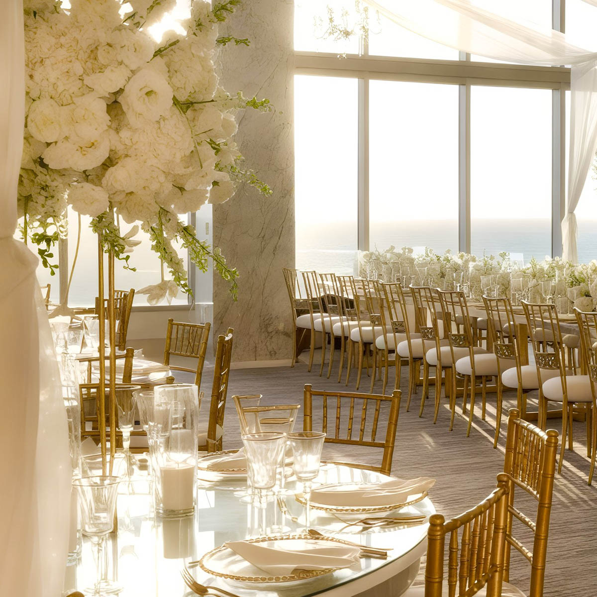 Elegant wedding reception setup with gold chairs, white floral arrangements, and dinnerware on tables in a sunlit room with large windows overlooking the ocean.