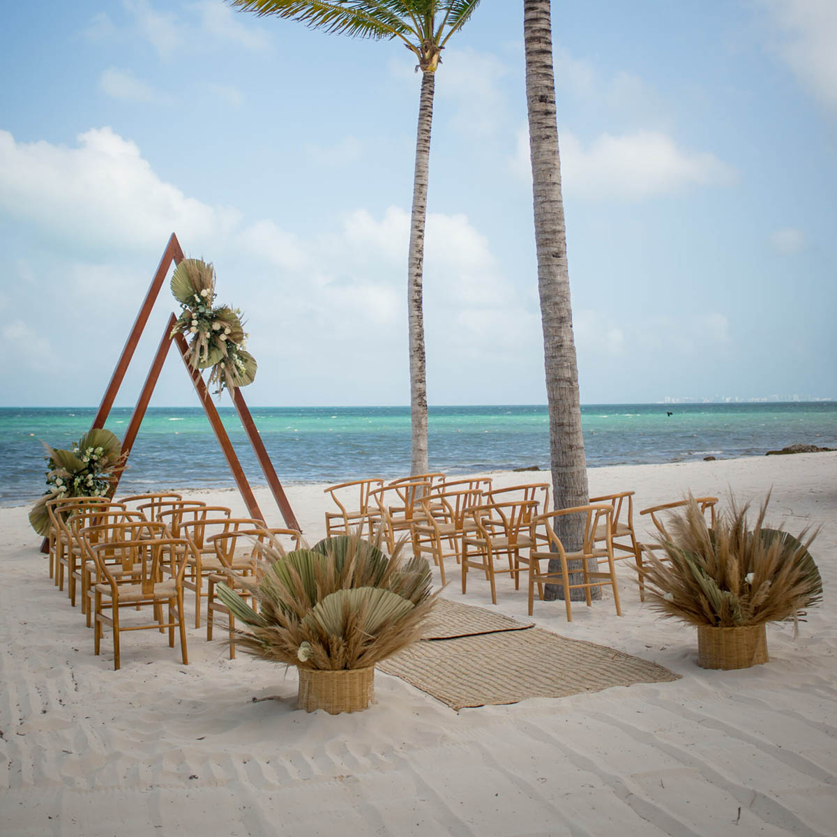 Beach wedding setup with wooden chairs facing a triangular arch decorated with dried leaves. Located on sandy shore with palm trees and ocean in the background.