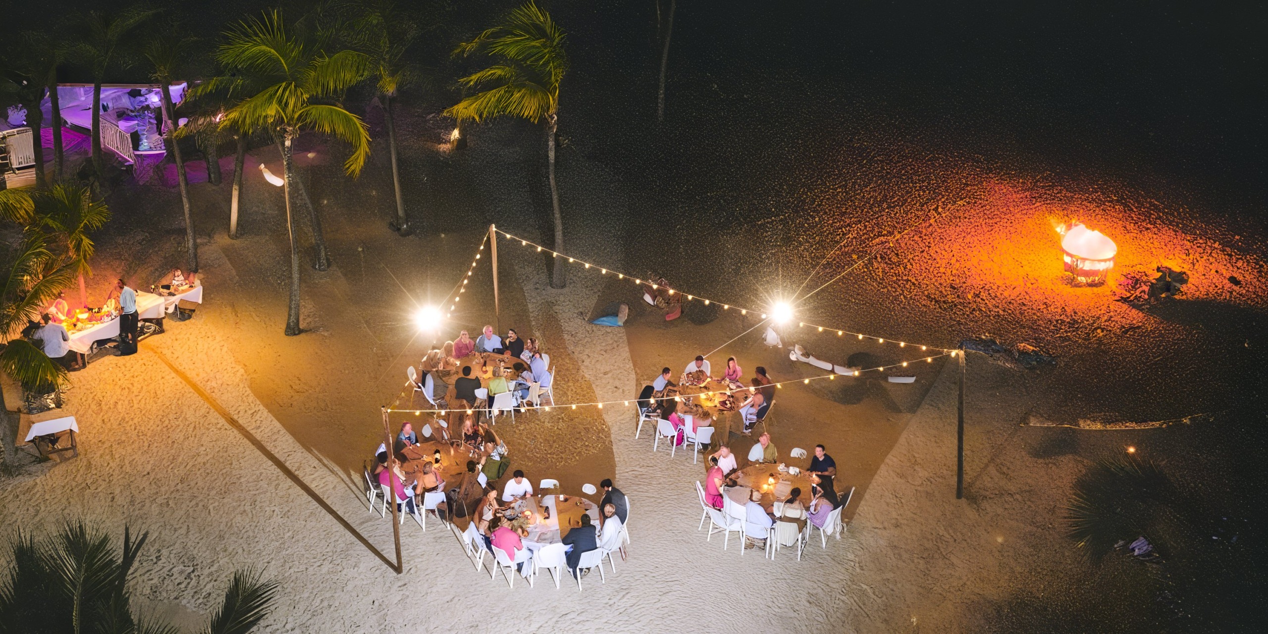 An outdoor beach dinner at Alexandra Resort Turks and Caicos features guests seated at round tables under string lights with a nearby bonfire surrounded by palm trees.