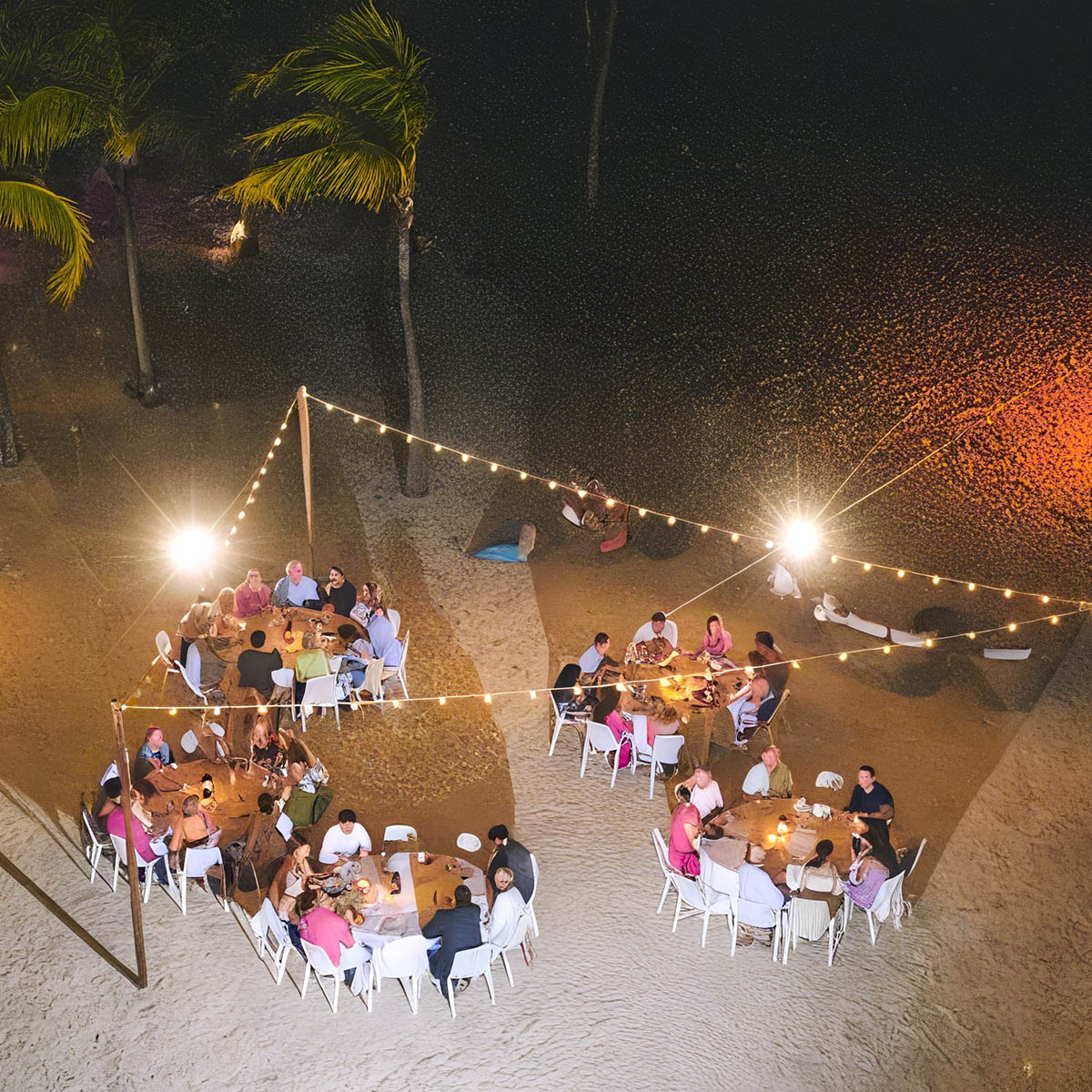 Aerial view of people dining at three tables on a sandy beach, decorated with string lights and surrounded by palm trees.