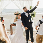 A couple in wedding attire stands under a canopy on a beach, holding hands. The groom waves while the bride holds a bouquet.