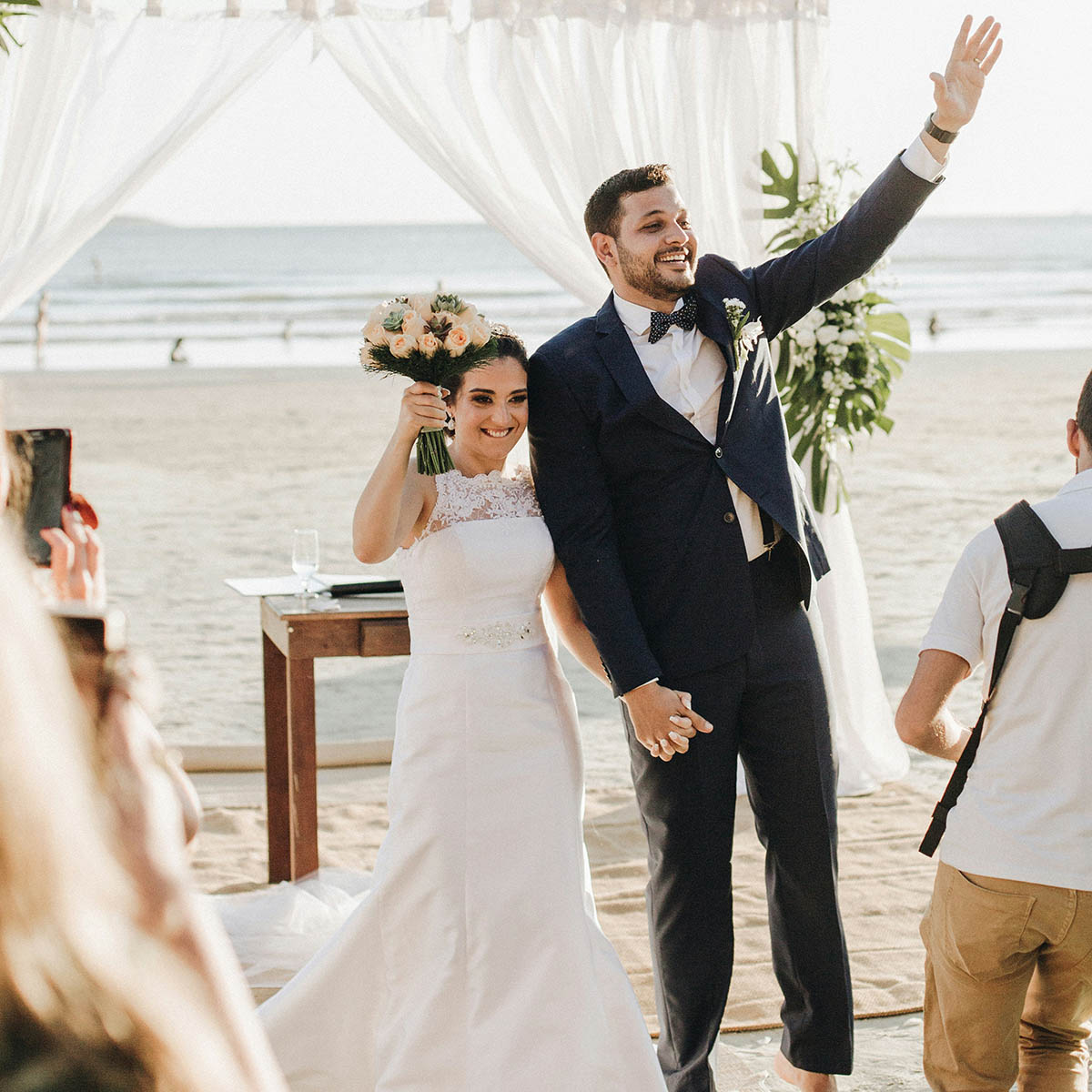 A couple in wedding attire stands under a canopy on a beach, holding hands. The groom waves while the bride holds a bouquet.