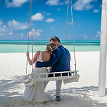 A couple, back to the camera, sits on a swing at a beach, looking at the turquoise ocean under a clear blue sky.