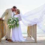 Bride and groom kiss on a wooden walkway by the beach. Bride holds a large bouquet and her veil flows in the wind.