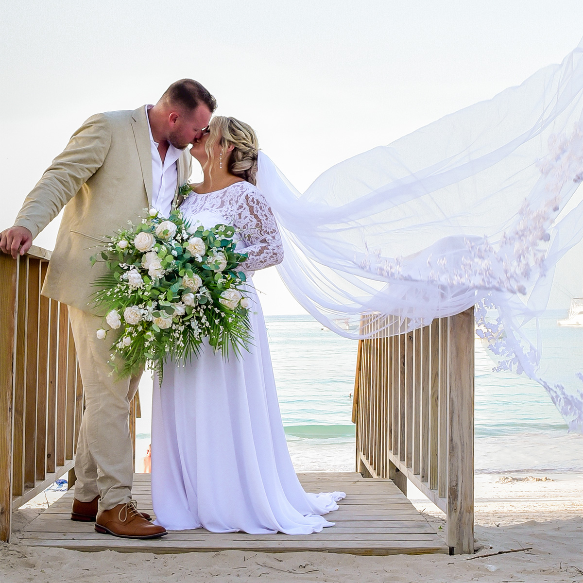 Bride and groom kiss on a wooden walkway by the beach. Bride holds a large bouquet and her veil flows in the wind.