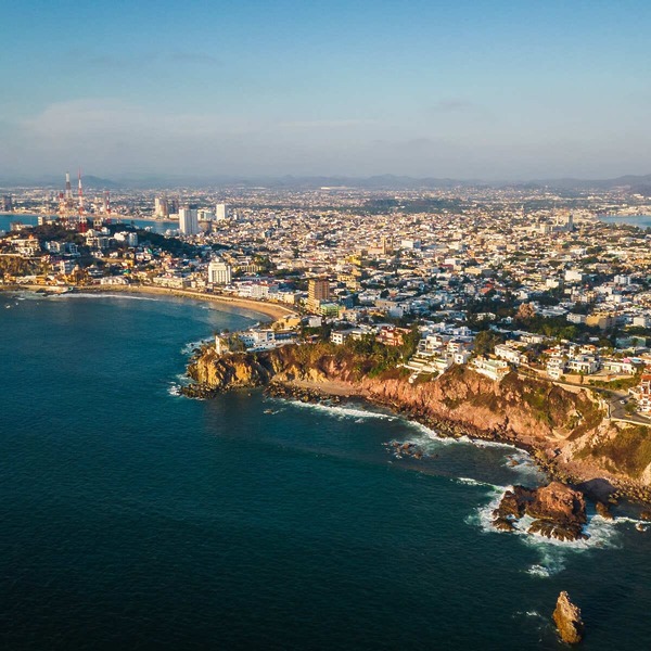 Aerial view of a coastal city with a rocky shoreline and urban landscape extending into the distance under a clear sky.
