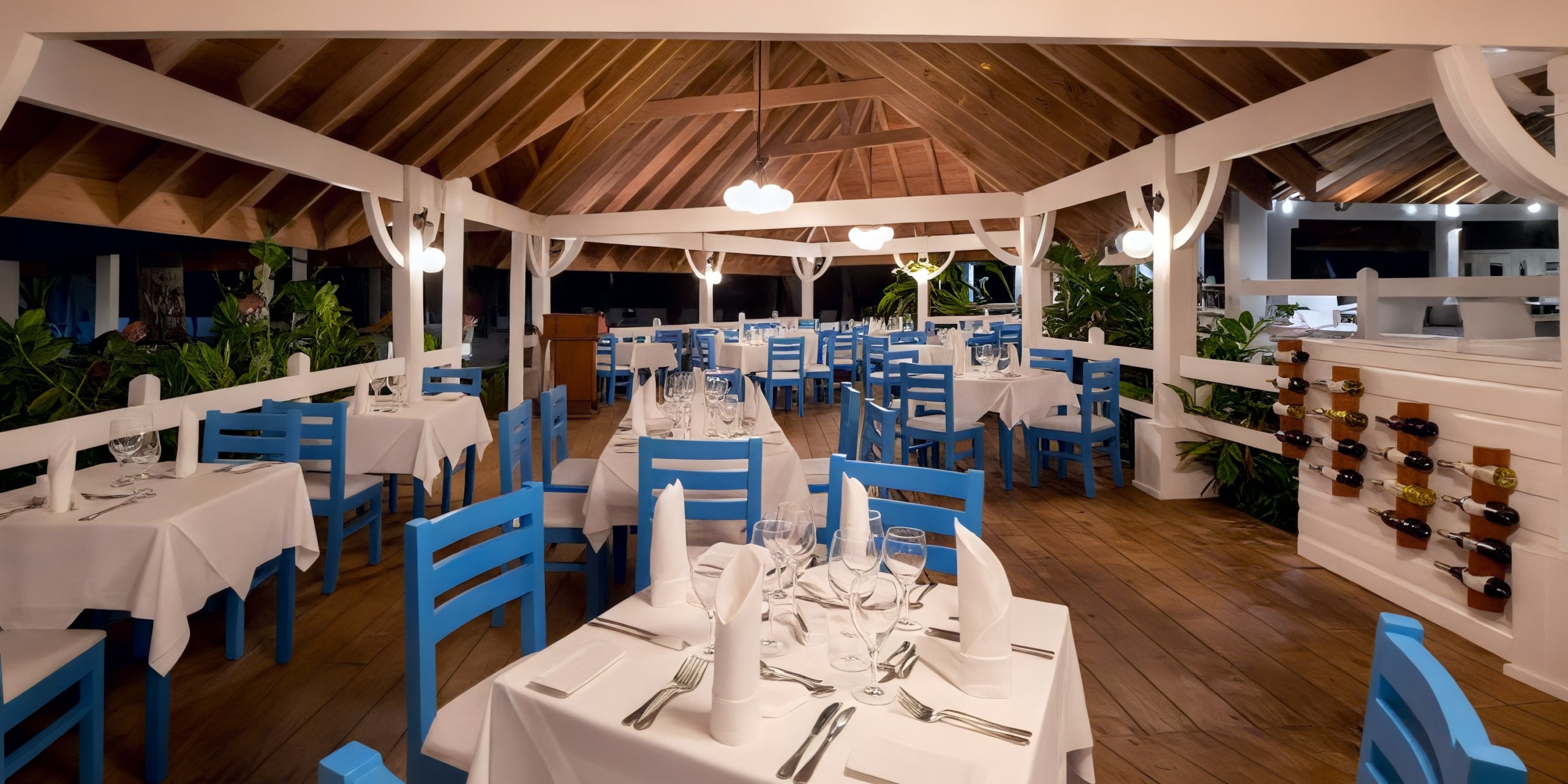 At Viva V Samana by Wyndham, the restaurant dining area features wooden ceilings, blue chairs, and white tablecloths. Tables are elegantly set with glassware and napkins.