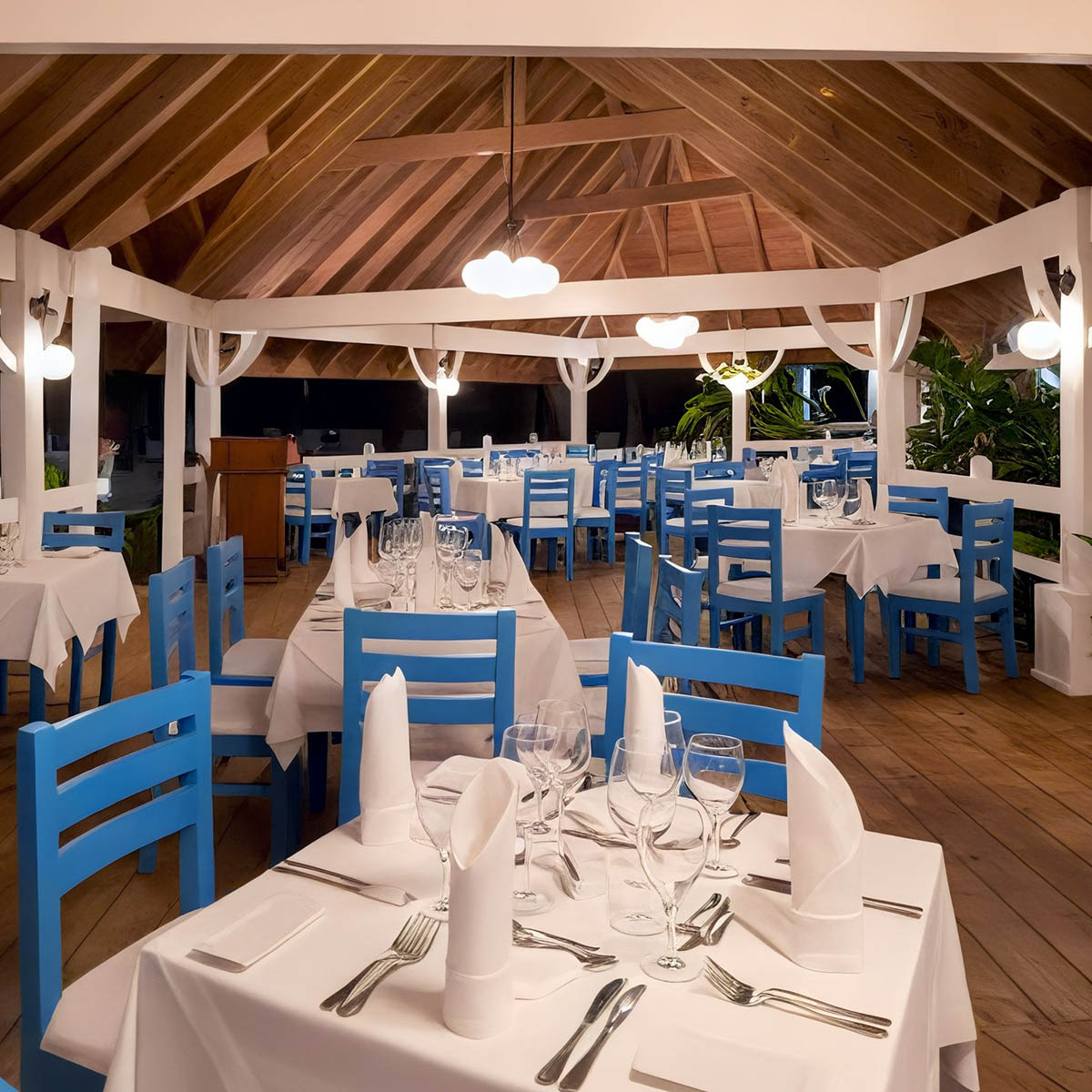 A dining area with blue wooden chairs and tables set with white tablecloths and napkins. The space has a wooden ceiling and ambient lighting.