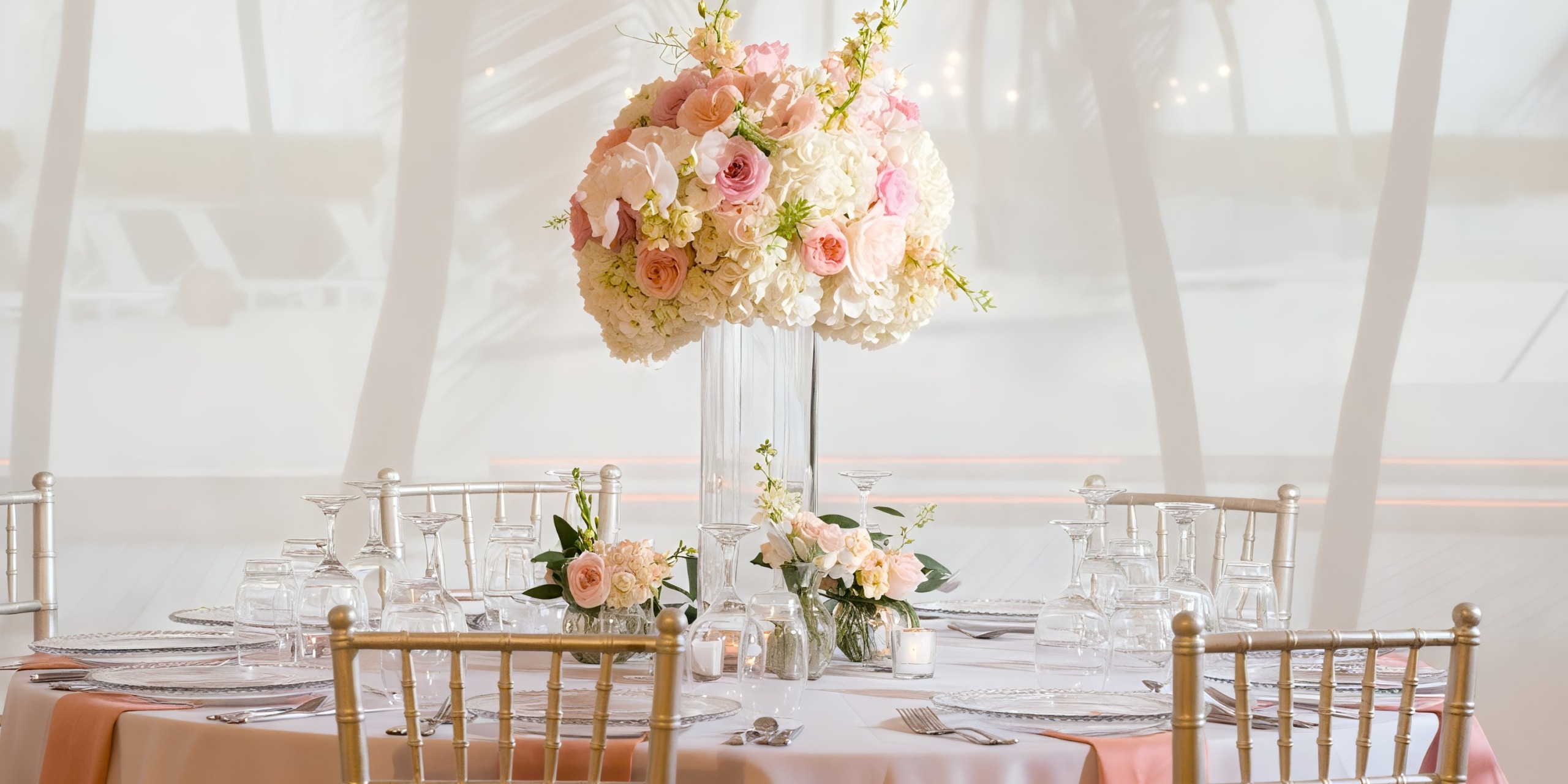 A sophisticated wedding table adorns the Alexandra Resort Turks and Caicos with a pink and white floral centerpiece, glassware, and white plates, set in an elegantly lit background.