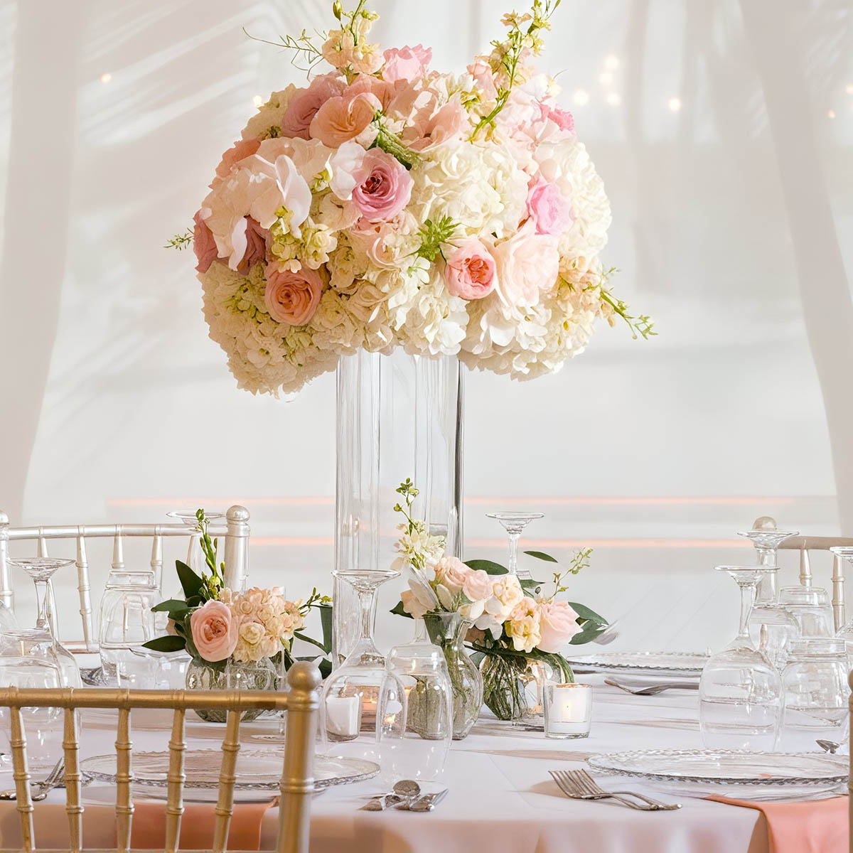 A tall floral centerpiece with pink and white flowers on a round table set with glassware and plates, surrounded by elegant chairs.