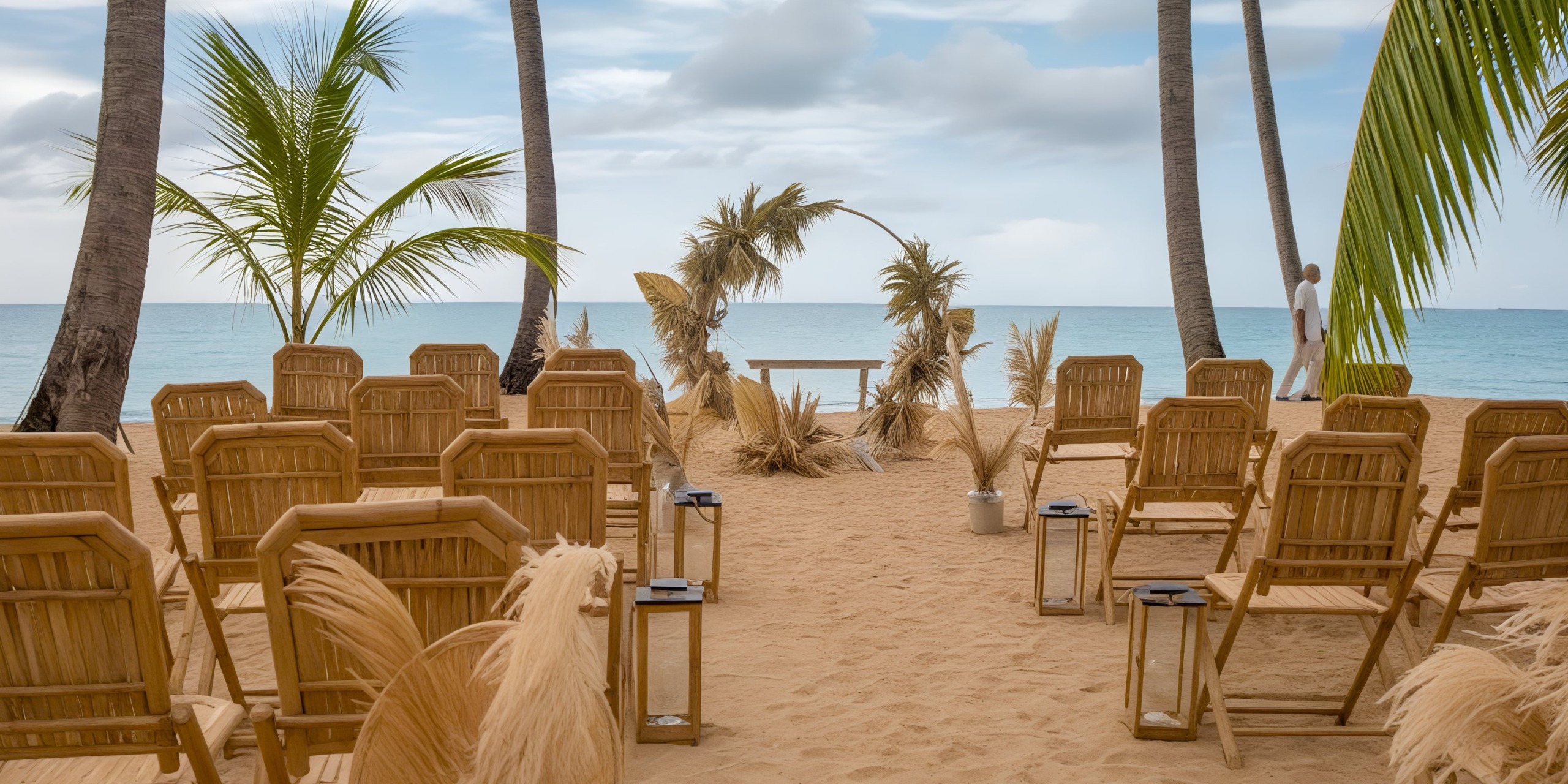 A tropical beach wedding setup at Viva V Samana by Wyndham features bamboo chairs, a small arch adorned with dried palm leaves, and the ocean as a backdrop under a cloudy sky.