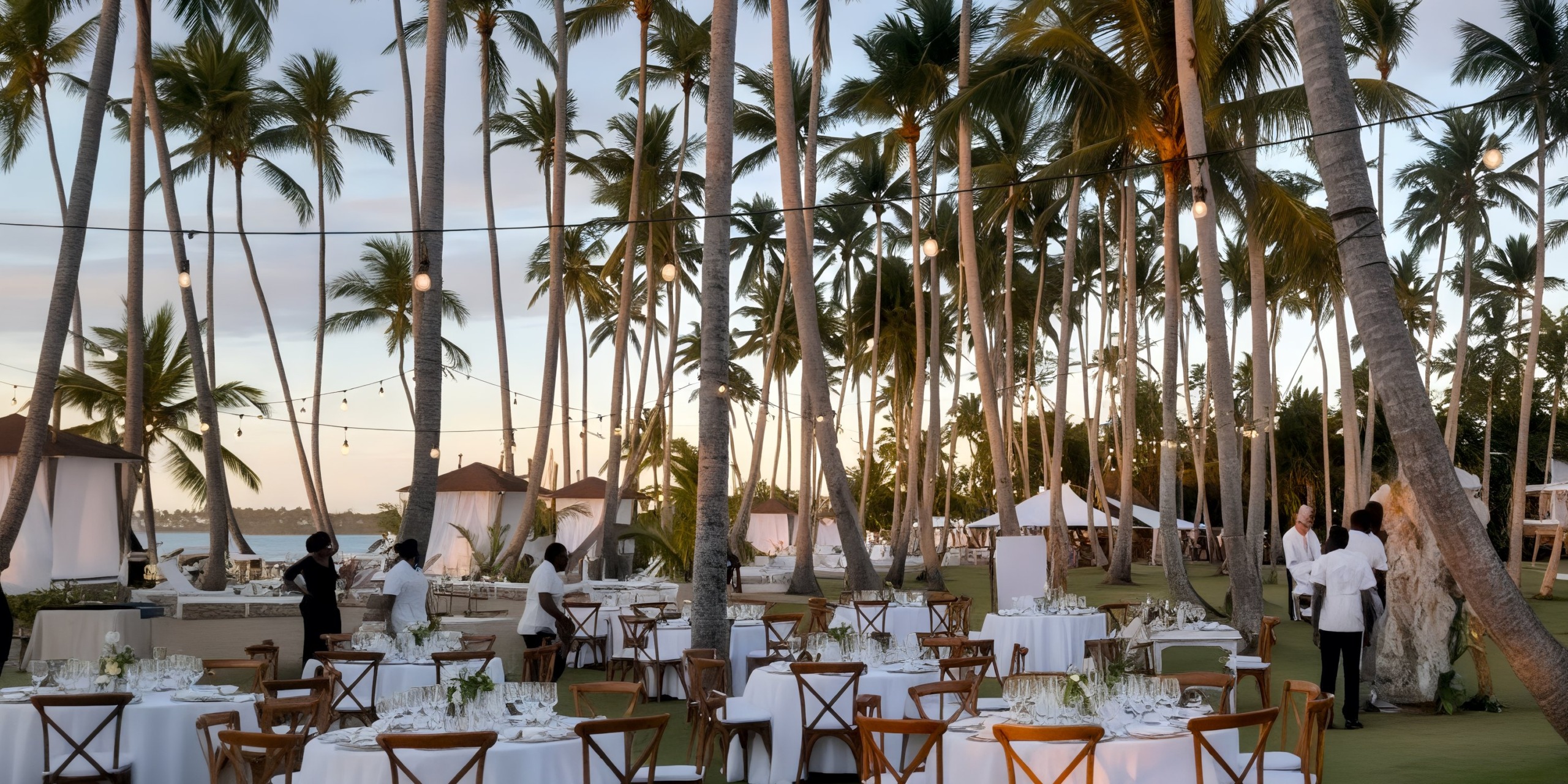 The outdoor dining area at Viva V Samana by Wyndham features tables and chairs on a grassy surface amid palm trees, with staff preparing the space under string lights as the sun sets.