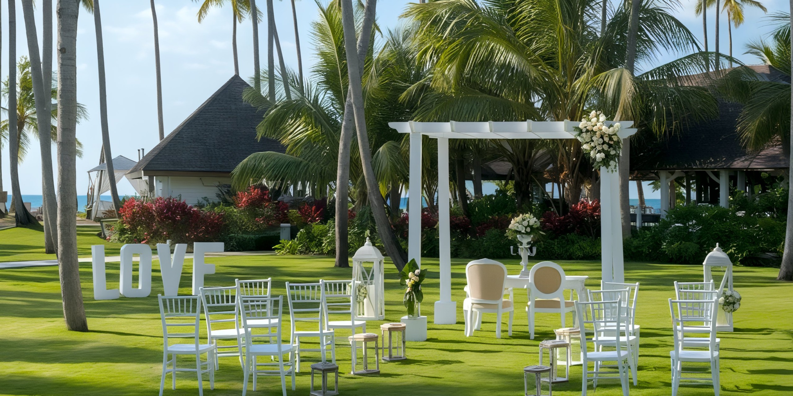 An outdoor wedding setup at Viva V Samana by Wyndham features white chairs and a small archway on a palm tree-surrounded lawn, with large "LOVE" letters displayed on the grass.