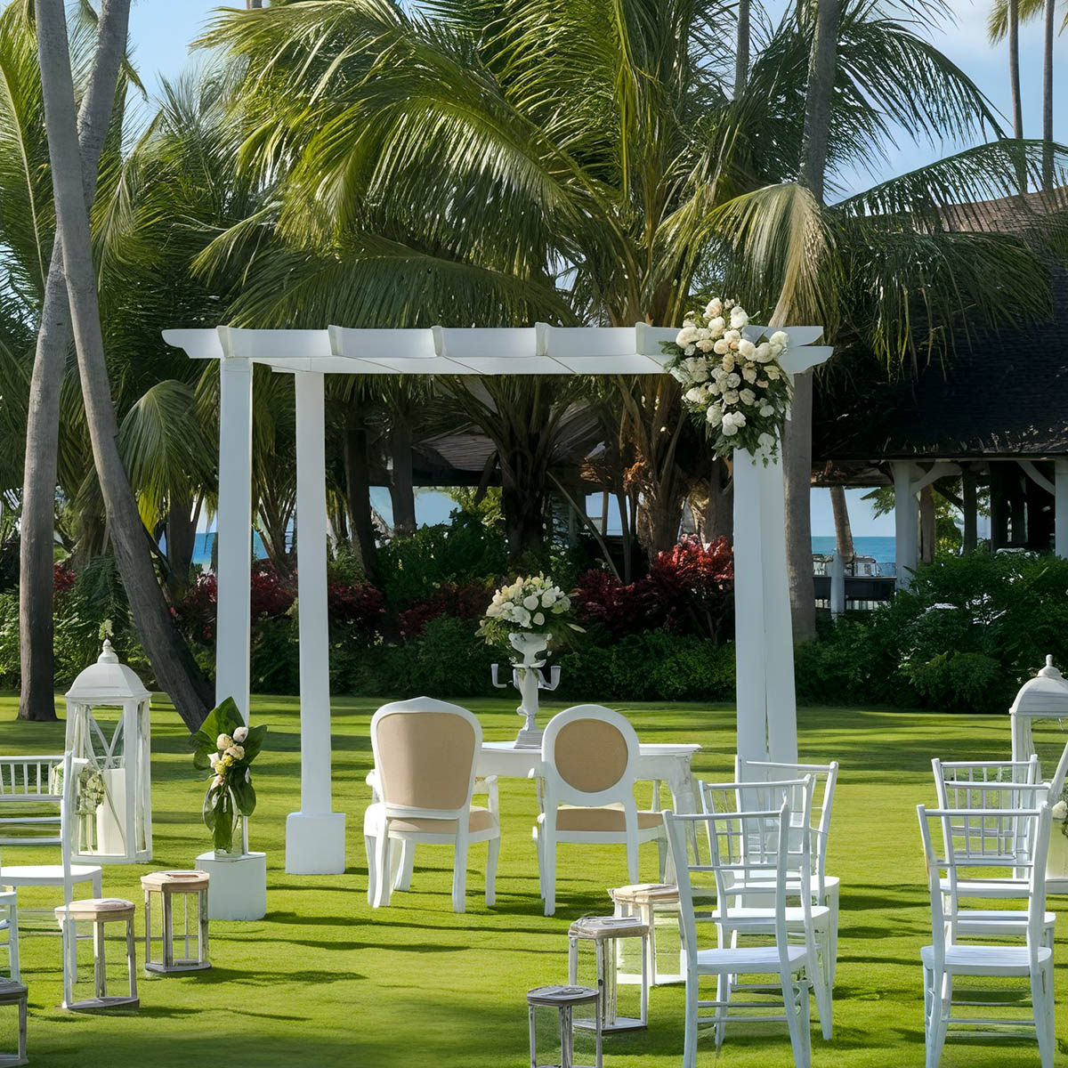 Outdoor wedding setup with a white arch adorned with flowers, surrounded by palm trees. White chairs and decorative lanterns are arranged on a green lawn.