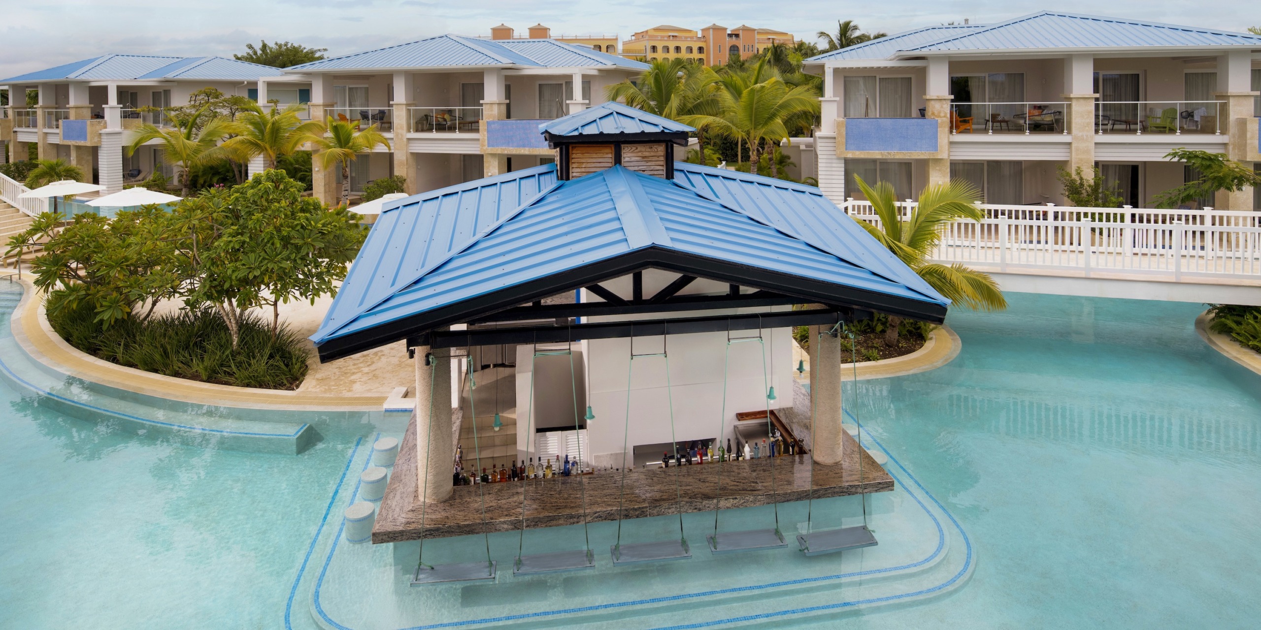 At Dreams Cap Cana Resort & Spa, enjoy a poolside bar with a blue roof in the center of the resort pool, surrounded by palm trees and resort buildings with blue accents.