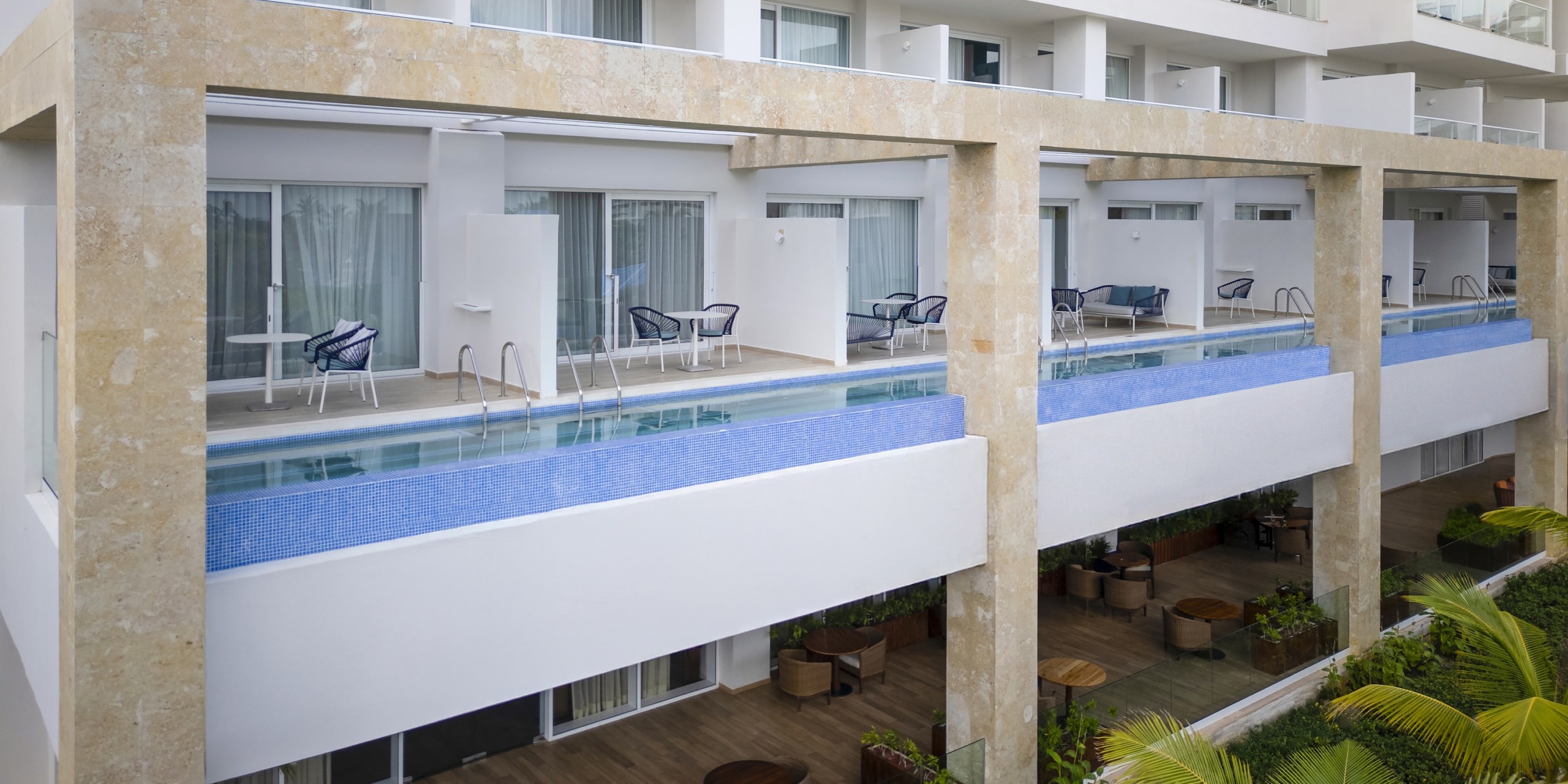Dreams Cap Cana Resort & Spa is a modern two-story hotel featuring balconies, and the upper floor offers a narrow pool lined along it with chairs and tables available on both levels.