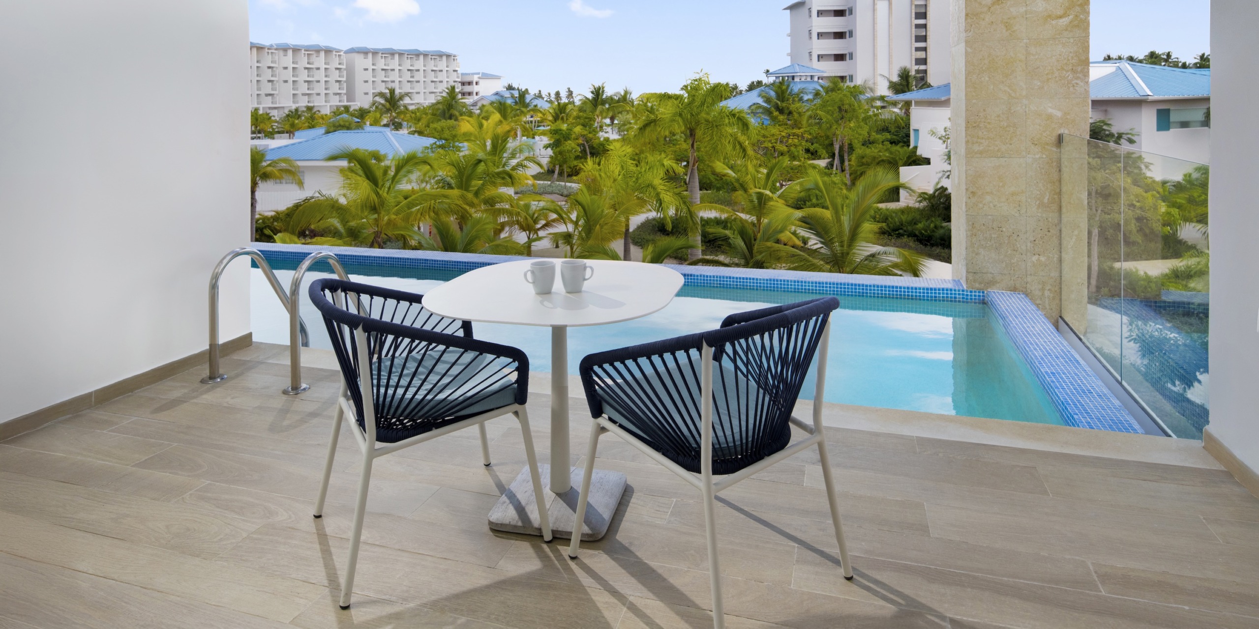 Two black chairs and a white table sit on a wooden deck beside the infinity pool at Dreams Cap Cana Resort & Spa, with views of lush tropical greenery and nearby buildings.