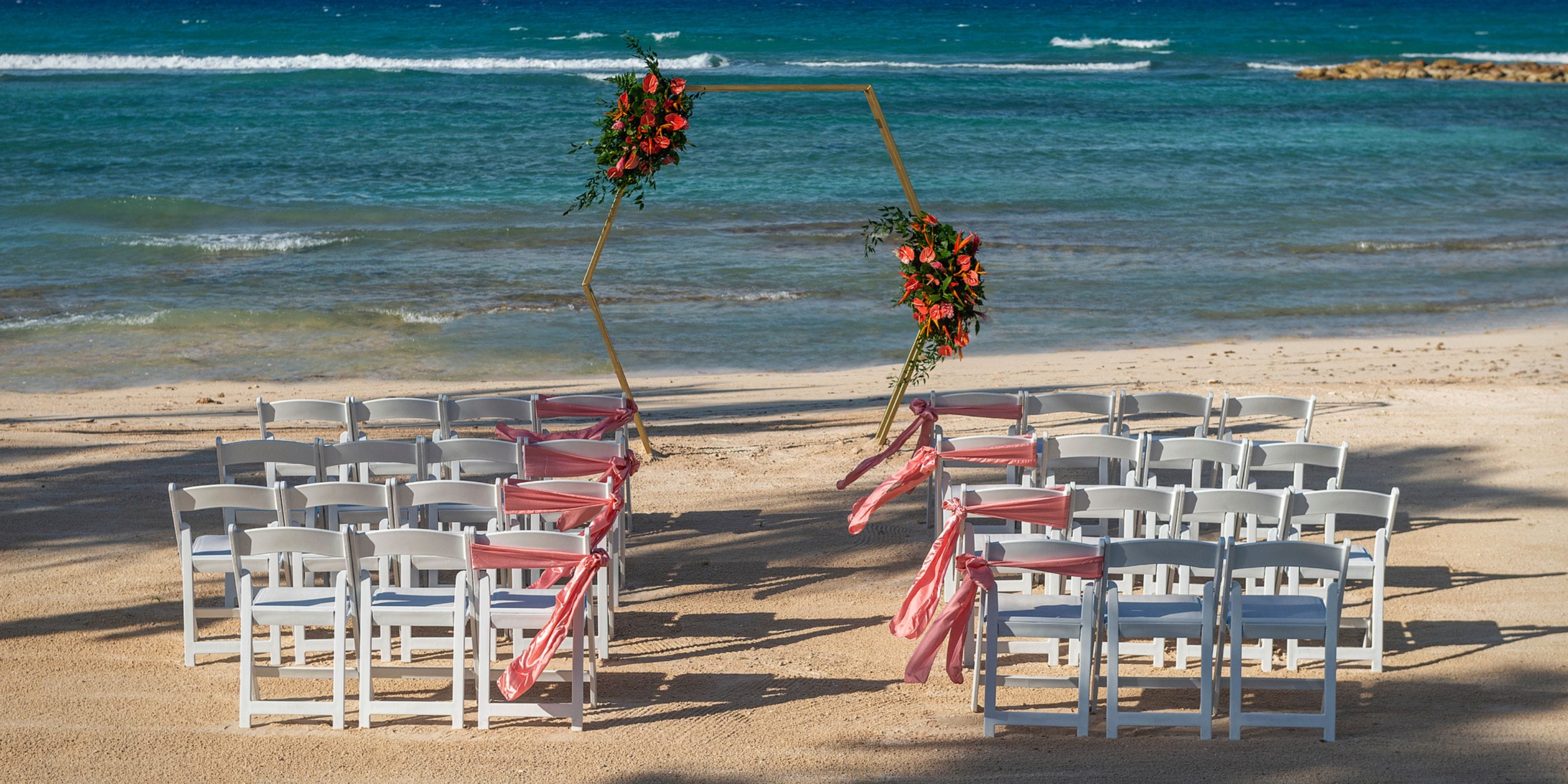 White chairs arranged for a destination wedding face a floral arch on the sandy beach, ocean in view.