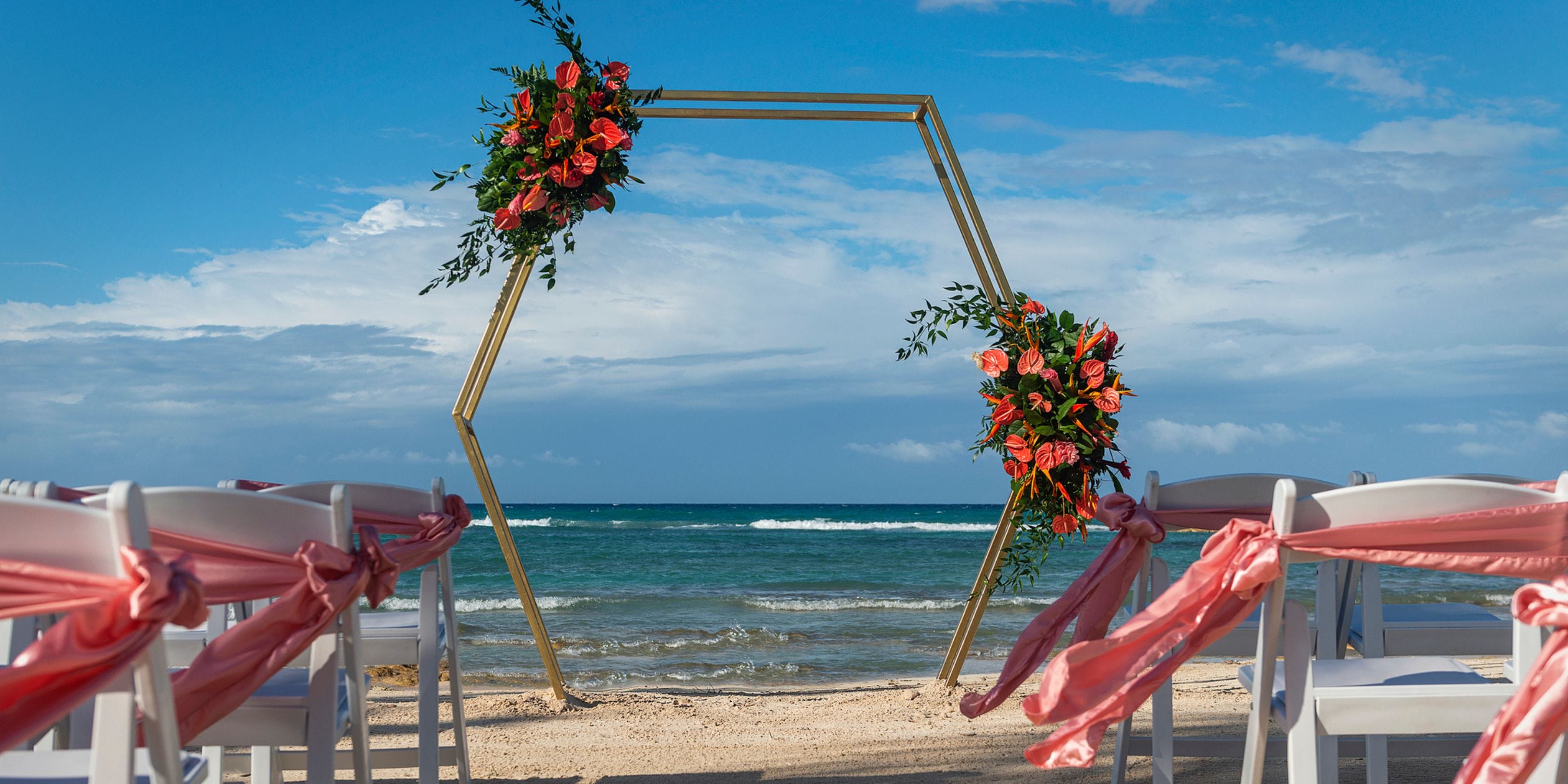 Hexagonal wedding arch with flowers on a beach at Ocean Eden Bay, pink-ribbon chairs for destination wedding.