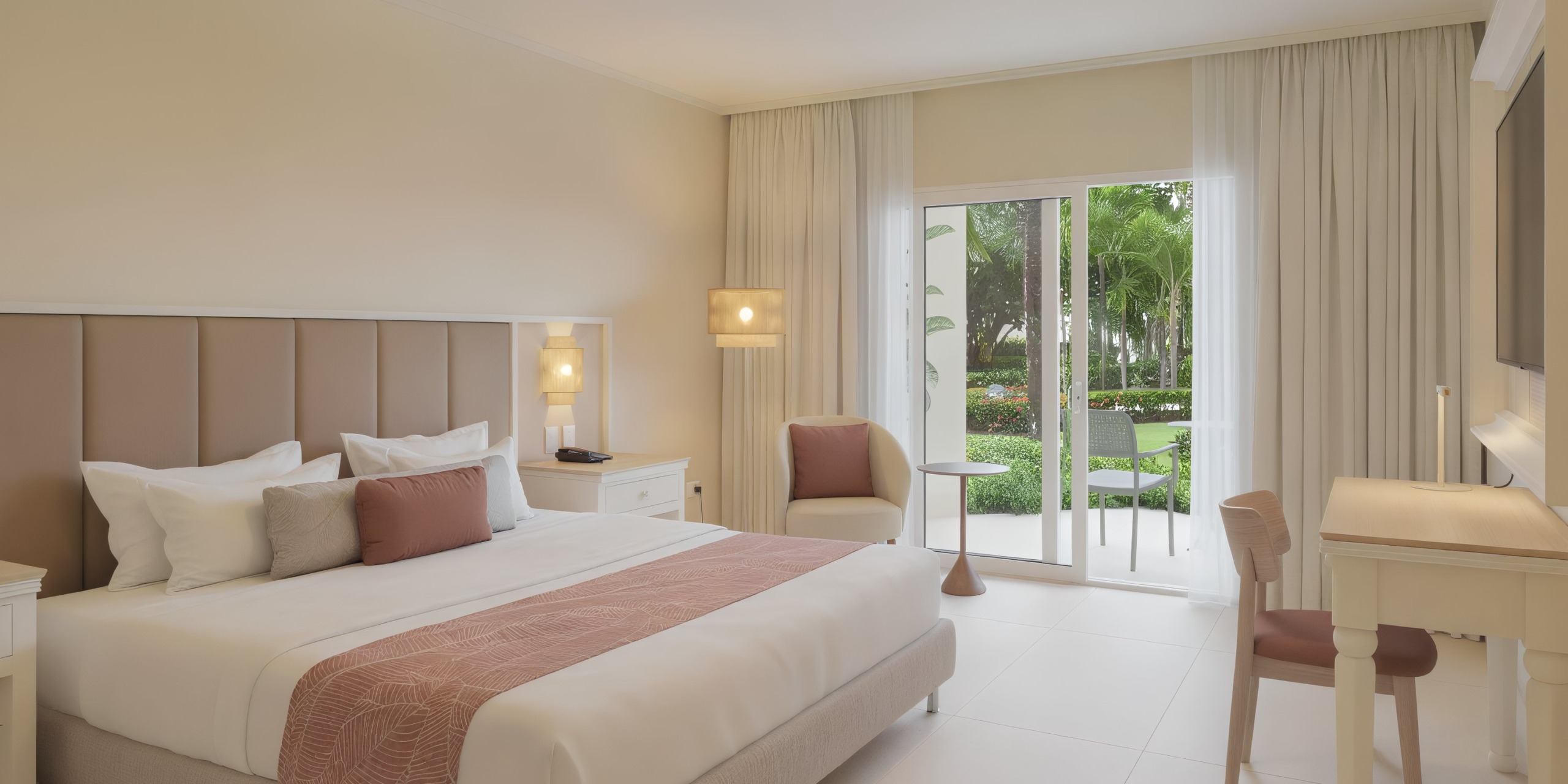 The Wyndham Alltra Punta Cana hotel room offers a neatly made double bed, two side tables with lamps, a desk and chair, plus a cozy seating area by the window with garden views.