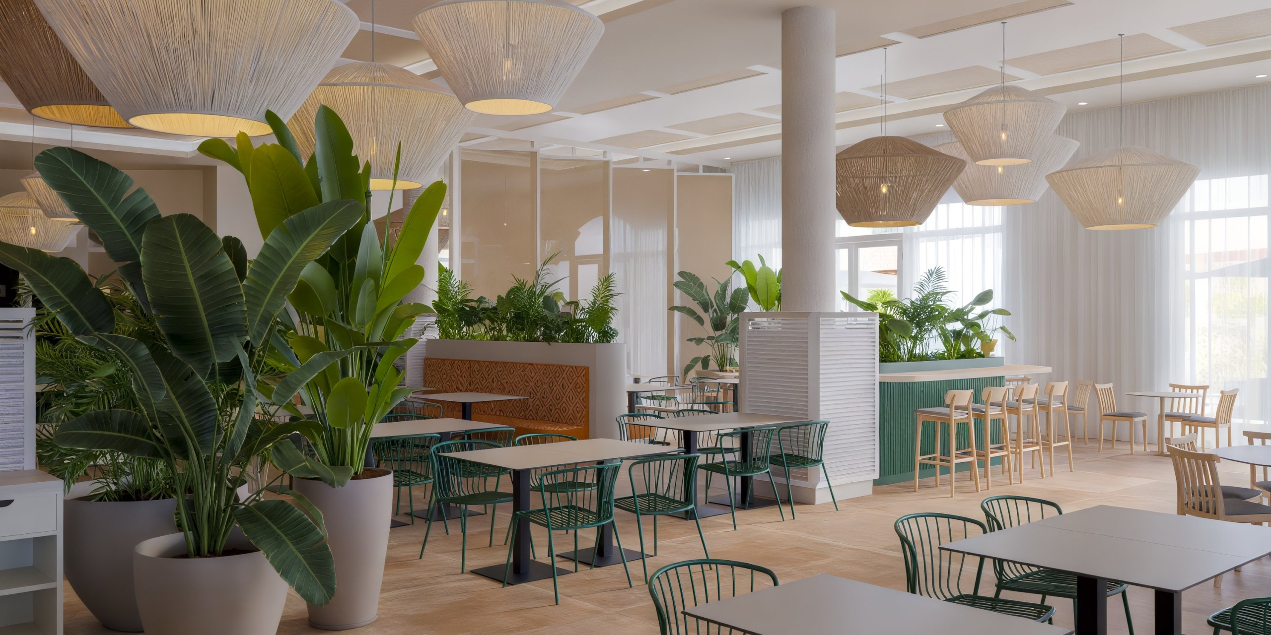 The Wyndham Alltra Punta Cana features a modern, airy cafe adorned with green plants and minimalist tables surrounded by green wire chairs and white hanging lights. Large windows flood the space with natural light.