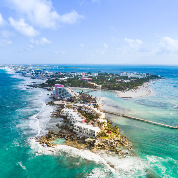 Aerial view of a coastal resort on a narrow peninsula with turquoise waters on both sides, sandy shores, and scattered buildings under a clear blue sky.