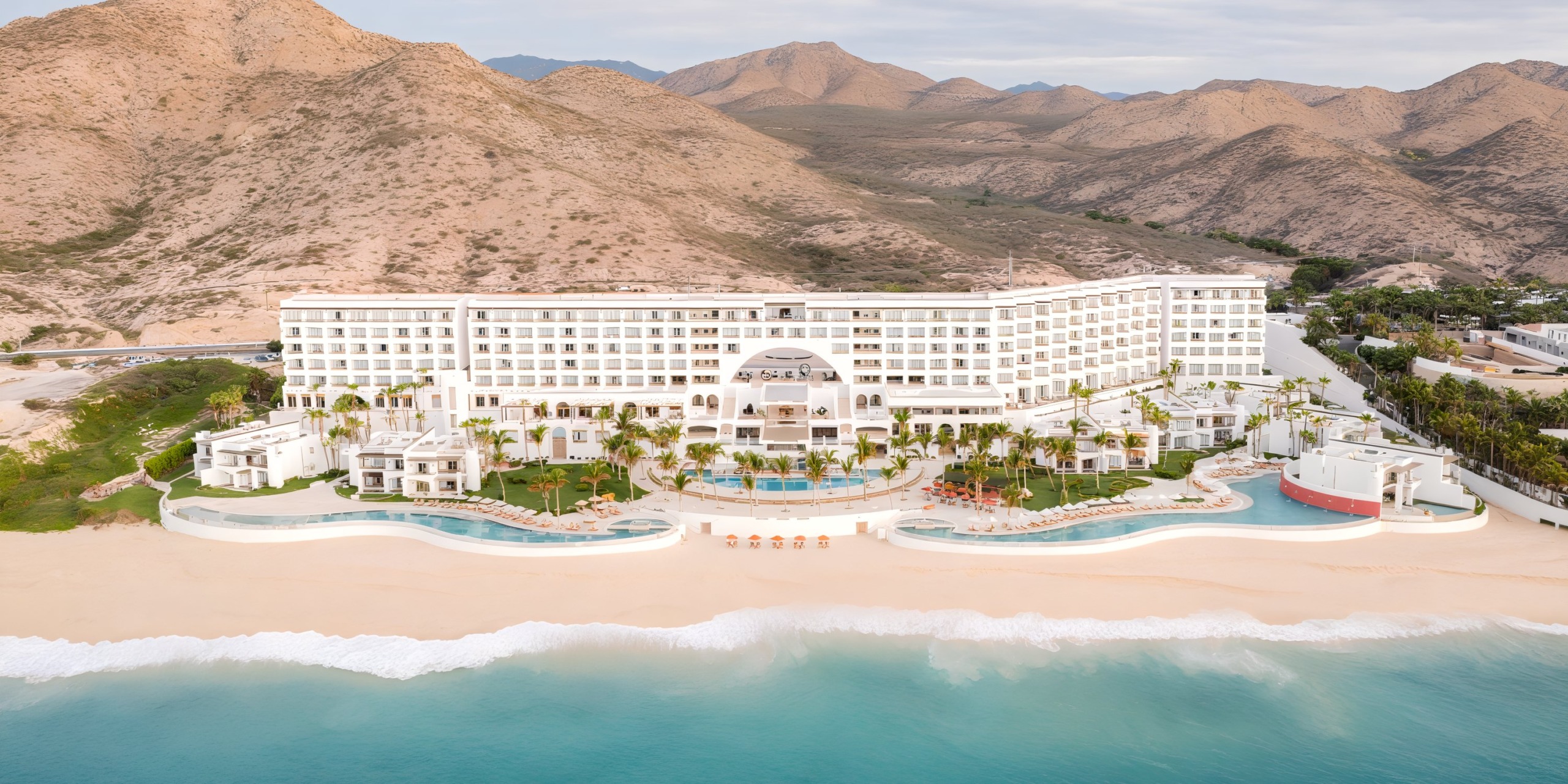 Aerial view of Marquis Los Cabos, a large beachfront resort featuring a U-shaped layout, multiple pools, palm trees, and surrounding mountains.