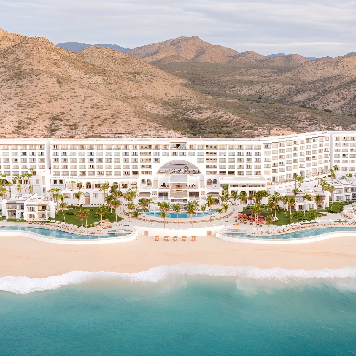 Aerial view of Marquis Los Cabos, a large beachfront hotel with swimming pools, surrounded by mountains and overlooking the ocean.